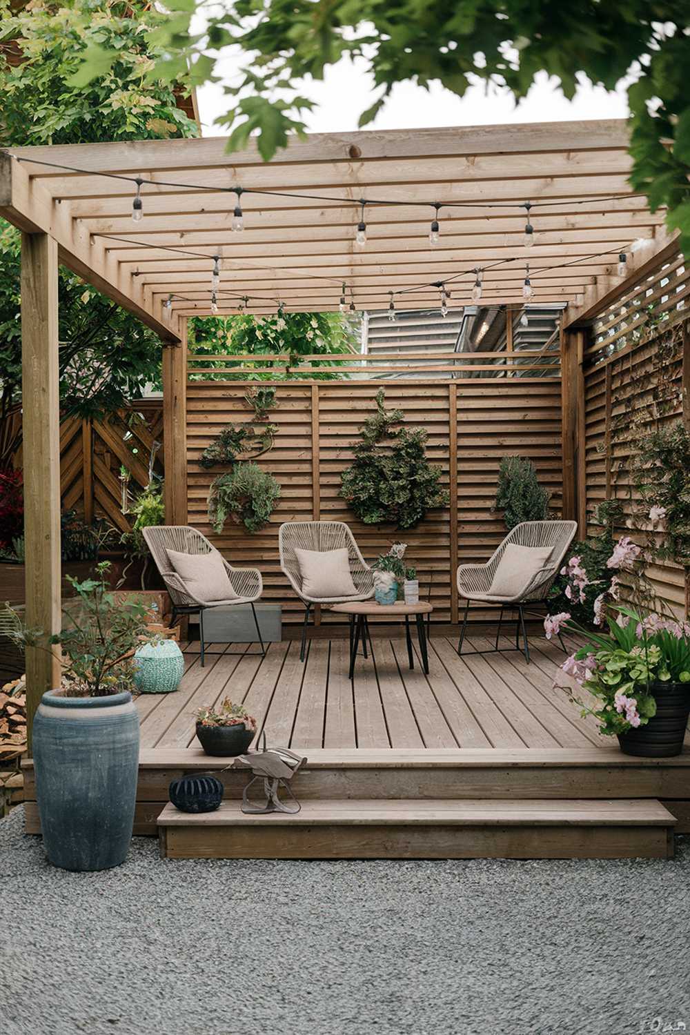 A backyard patio design and decor. There is a wooden deck with a few chairs and a small table. A few potted plants are placed around the deck. The deck is partially covered by a wooden pergola. There are string lights hung across the pergola. The ground is covered with gravel.