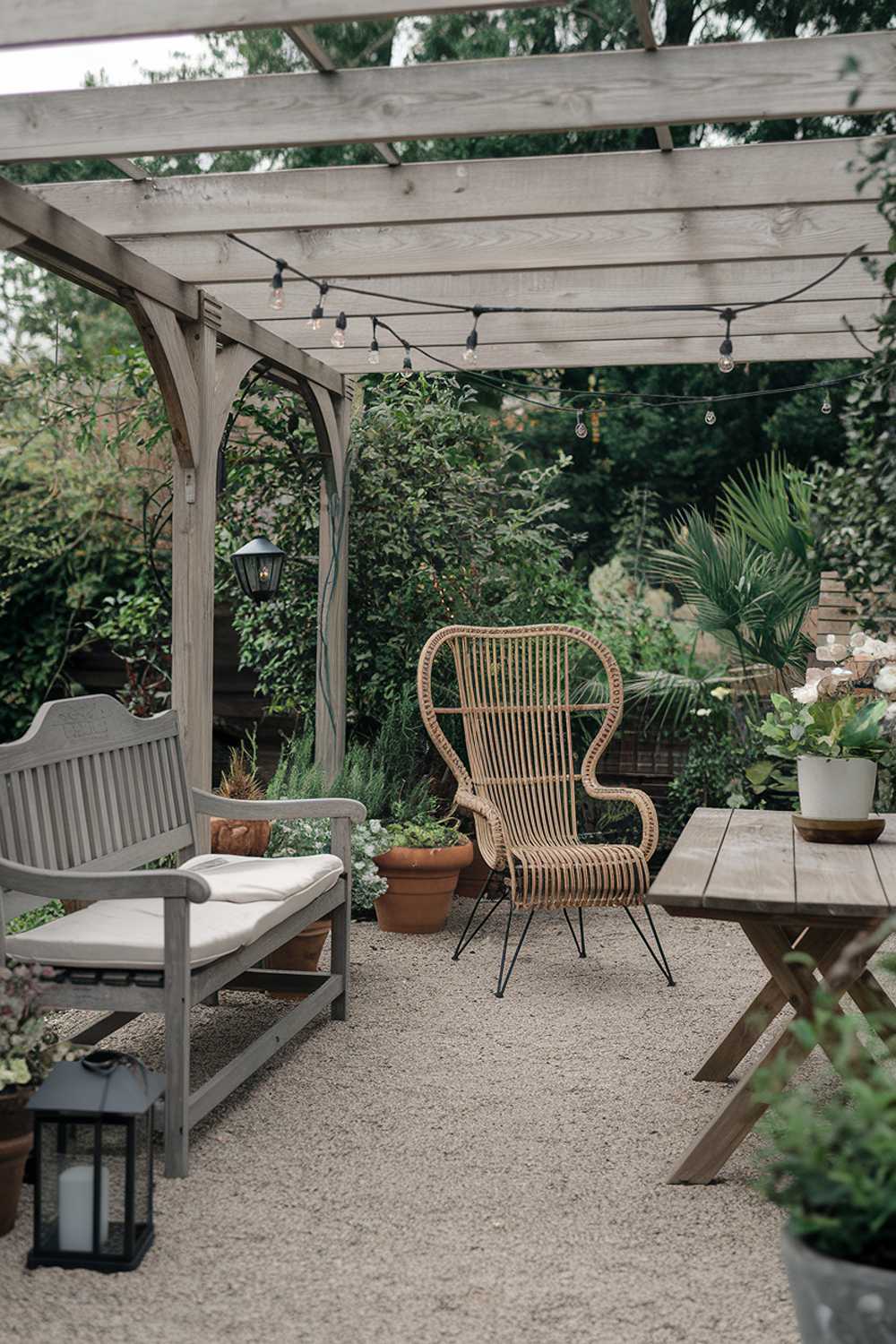 A backyard patio with a few pieces of furniture. There's a wooden bench with a backrest, a rattan chair, and a wooden table. The patio is covered with a wooden pergola. There are potted plants, string lights, and a lantern. The ground is covered with gravel. The background is lush with greenery.