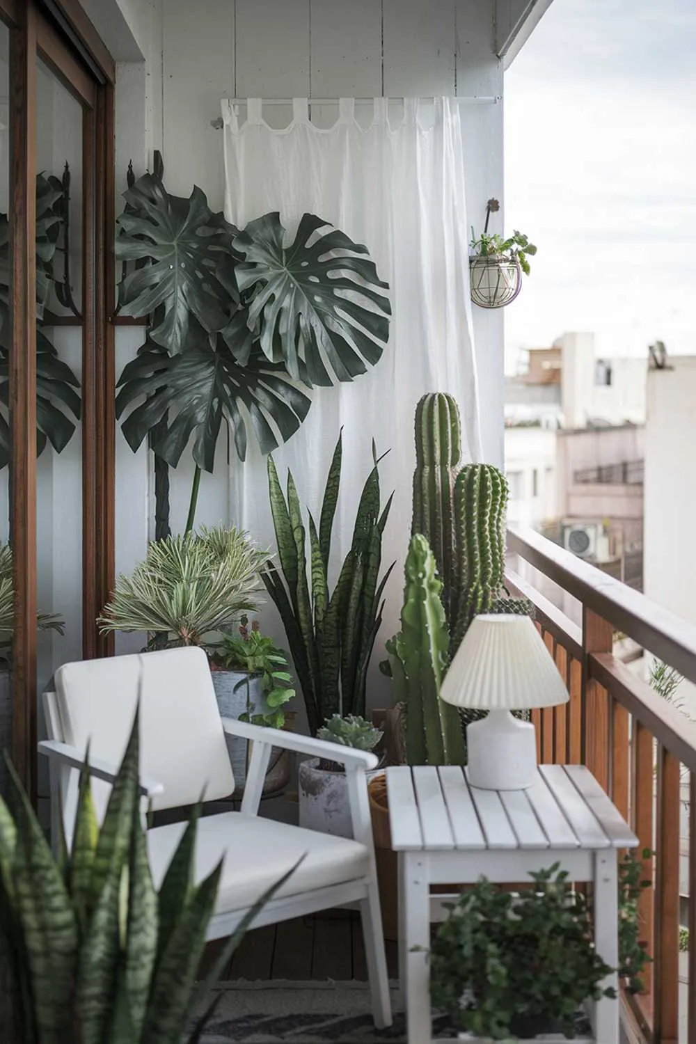 23 Perfect Balcony Plants Ideas for Small Spaces 2 A balcony with plants design and decor idea. There is a white wooden chair and a white wooden table on the balcony. There are various plants, including a large monstera plant, a snake plant, and a cactus. There is a white lampshade on the table. There is a white curtain on the wall. The balcony has a wooden railing. The background reveals a cityscape with buildings.