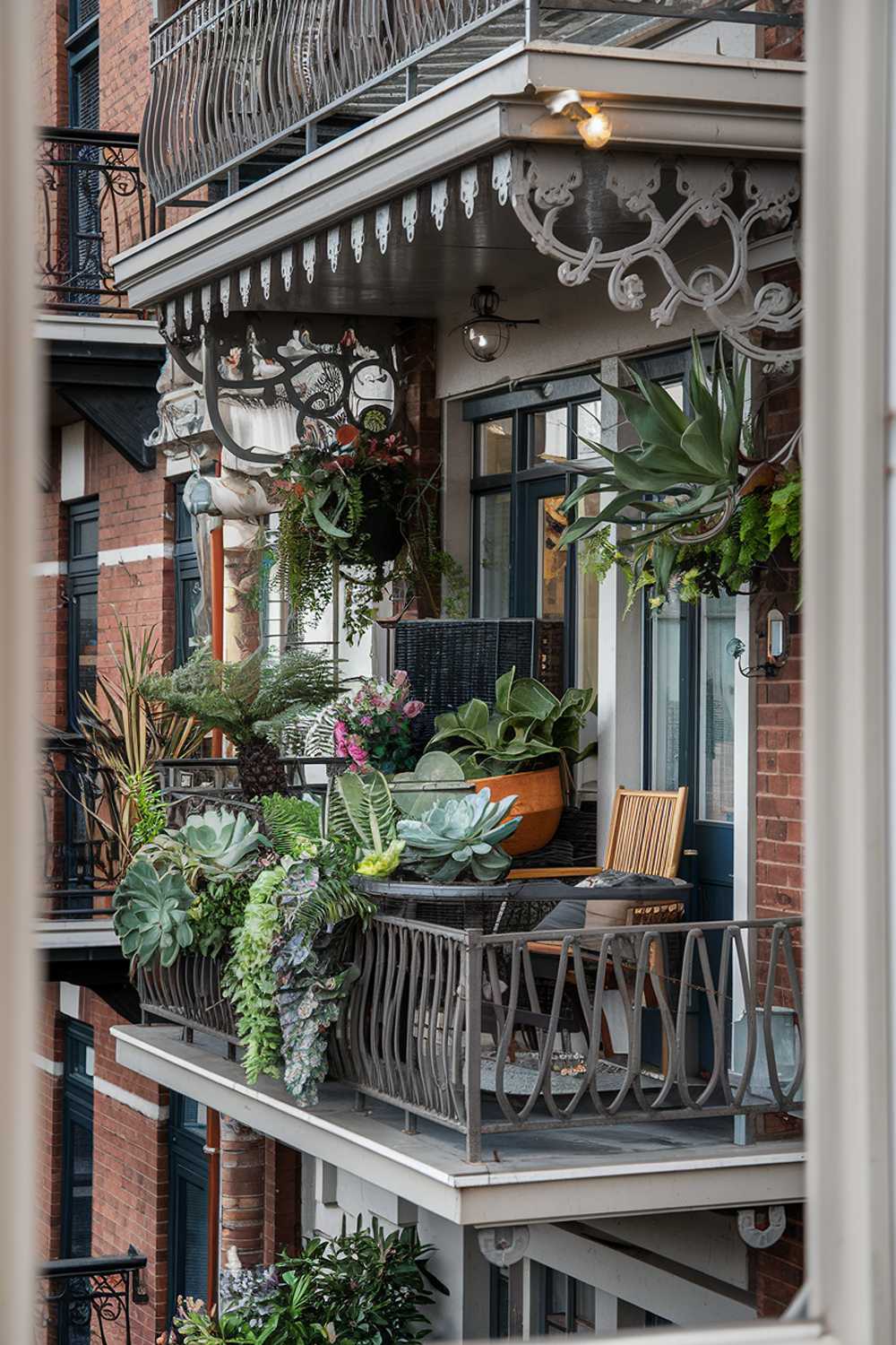 The balcony is decorated with various plants, including succulents, ferns, and flowers. There's also a wooden chair and a small table on the balcony. The railing of the balcony is made of metal and has a decorative pattern. The background contains a brick building with multiple balconies.