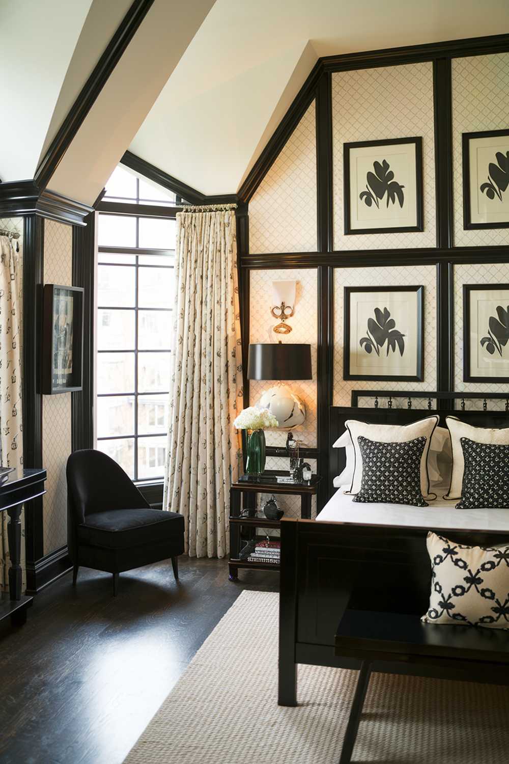 A black and cream bedroom with a stylish design. The room has a high ceiling and is illuminated by a large window. There's a black bed with cream pillows and a black lampshade on the bedside table. The walls have cream patterned wallpaper and black frames. There's a black chair near the window. The floor is made of dark hardwood.