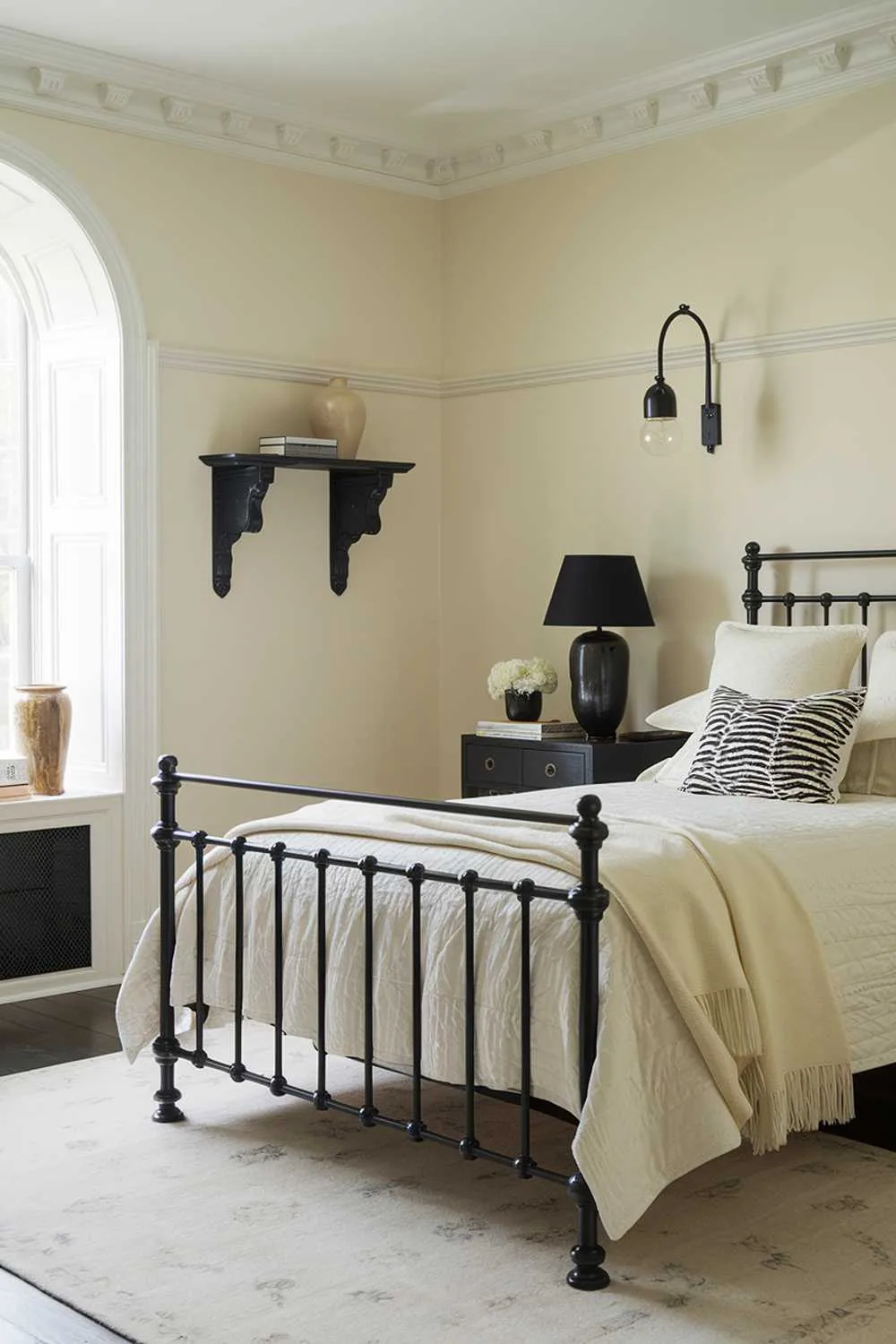 A black and cream bedroom design. The room features a black metal bed frame with cream bedding and a black wooden nightstand. There is a black lamp on the nightstand and a cream throw blanket on the bed. The walls are painted cream, and there is a black wall shelf with a cream vase near the window. The floor is covered with a cream area rug.