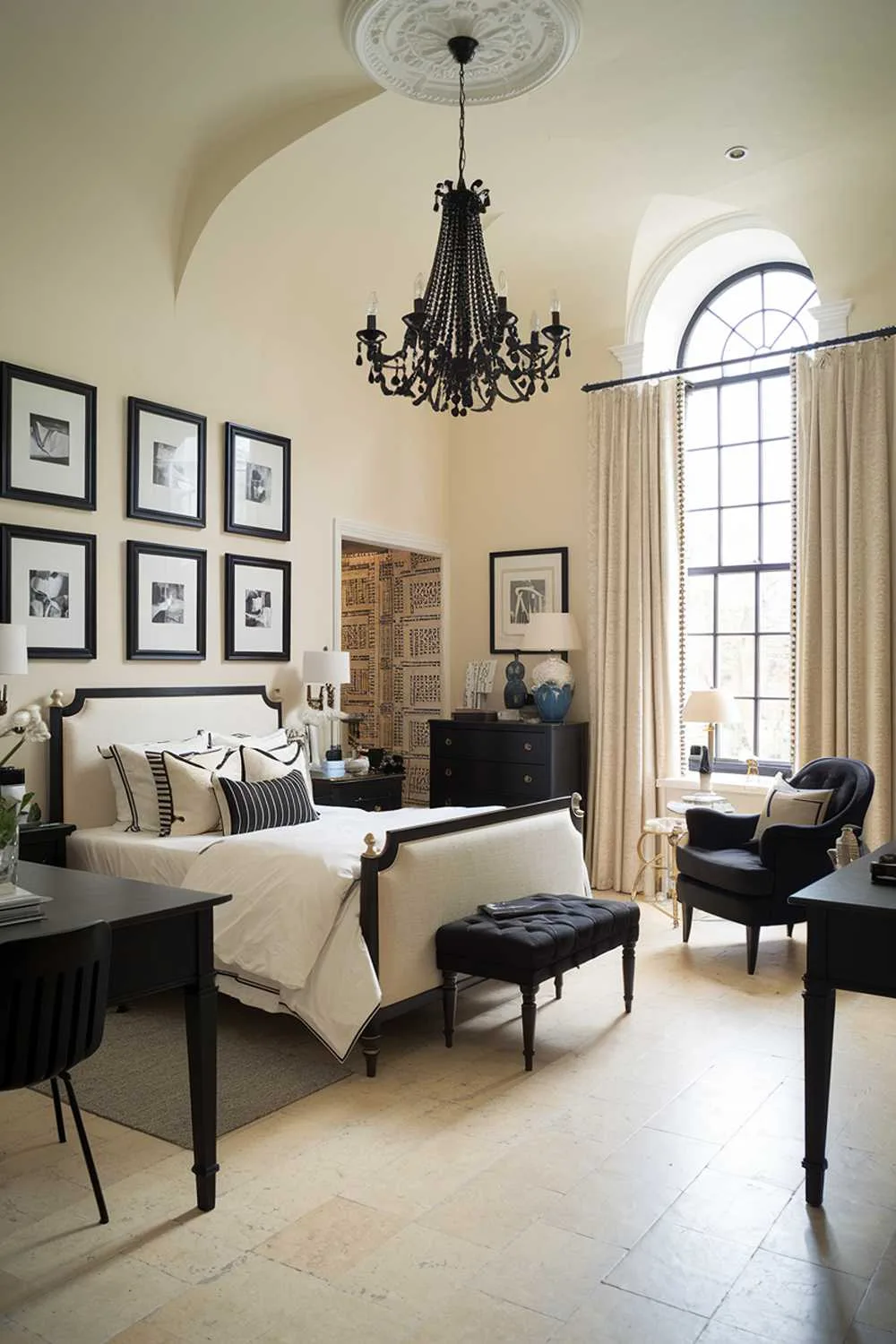 A black and cream bedroom design and decor. The room has a high ceiling, with a black chandelier hanging above the cream bed. The bed is placed against the wall, with a black headboard. There's a black dresser and a black desk in the room. The floor is made of cream tiles. The walls have black frames with white pictures. There's a black armchair near the window. The window has cream curtains.