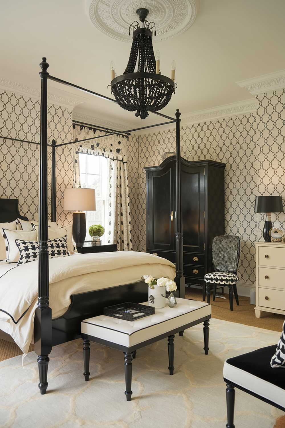 A black and cream bedroom design and decor. The room has a black four-poster bed with cream bedding. There's a black chandelier hanging above the bed. The walls are decorated with cream and black patterned wallpaper. The room has a black wardrobe, a cream dresser, and a black chair. The floor is covered with a cream area rug. The room has a cream lampshade and a black lamp.