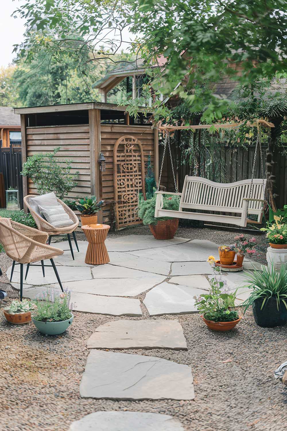 A backyard with a flagstone patio design. The patio is decorated with a variety of items, including a white porch swing, rattan chairs, a wooden side table, potted plants, and a decorative archway. There is a wooden structure in the background, possibly a shed or a storage area. The ground around the patio is covered with gravel, and there are plants in various pots. The background contains trees and greenery.