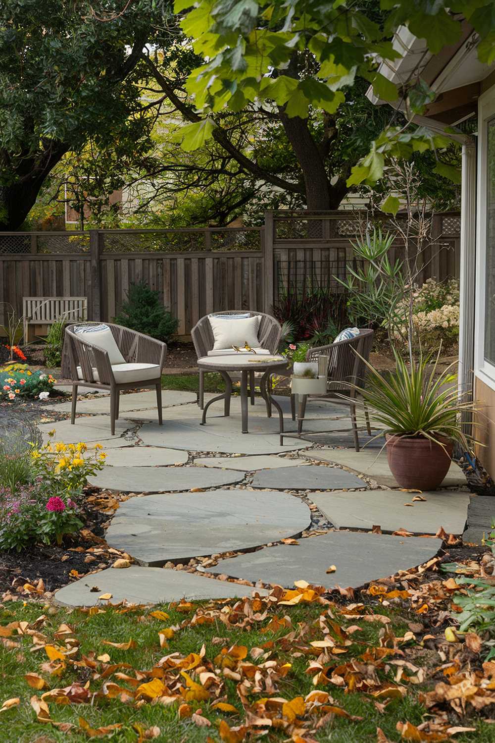 A backyard with a cozy flagstone patio design. The patio has a few chairs and a table. There's a potted plant next to the table. The background contains a wooden fence and lush greenery. The patio is surrounded by a garden with flowers and plants. The ground is covered with fallen leaves.