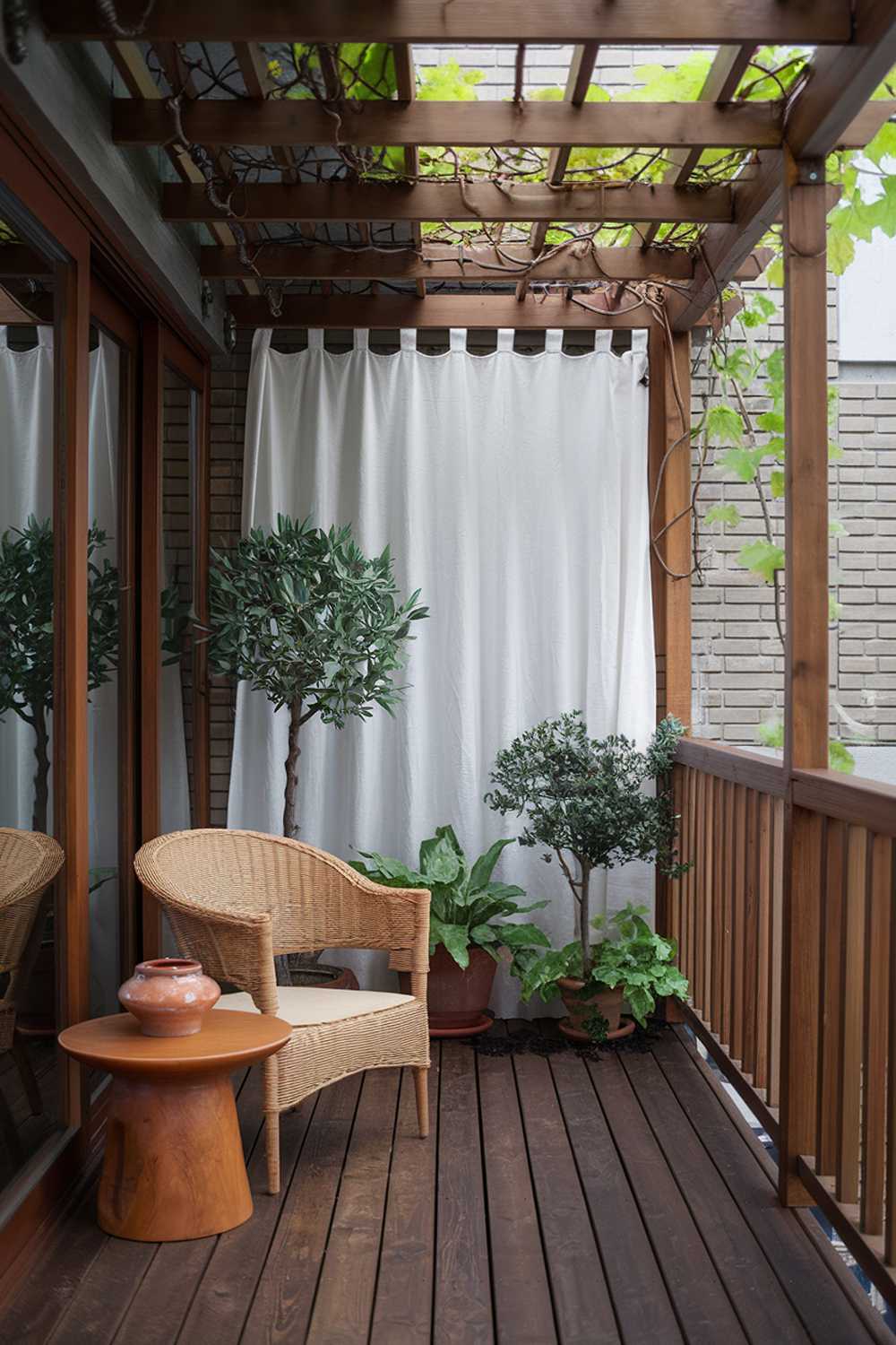 A Japandi balcony design and decor. The balcony features a wooden floor and a wooden pergola with vines growing on it. There are a few potted plants, including a small tree. A wicker chair and a wooden side table with a ceramic pot are placed on the floor. There is a white curtain attached to the wall. The railing is made of wood. The background contains a brick wall.