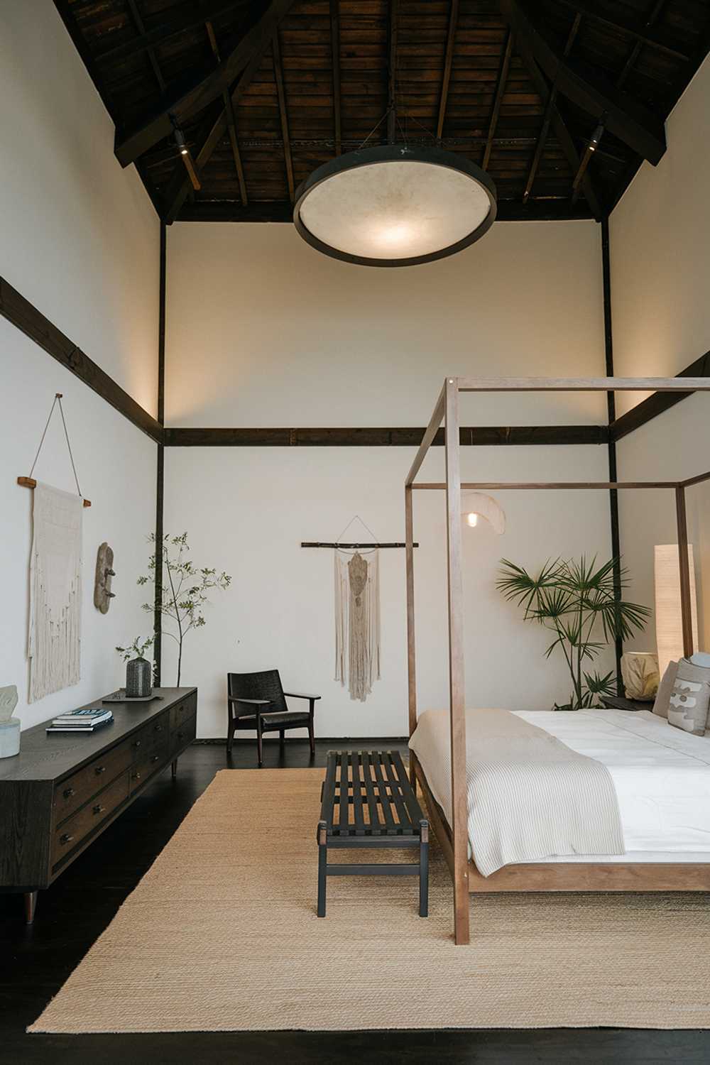 A Japandi-style bedroom with a high ceiling. The room has a king-size bed with a wooden frame. There's a beige rug beneath the bed. A long, dark wooden dresser is placed against one wall, holding a few items. A dark wooden chair is placed near the bed. The walls have a few hanging items. There's a potted plant near the dresser. The room is lit by a large, circular light fixture hanging from the ceiling.