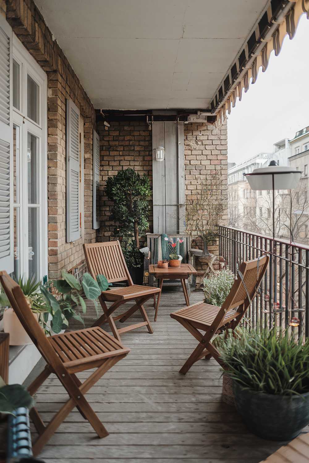 A large balcony with a rustic design. There are wooden chairs, a table, and a plant. The balcony has a brick wall and a railing. The floor is made of weathered wood. There are potted plants and a lamp on the balcony. The background reveals a cityscape with buildings.