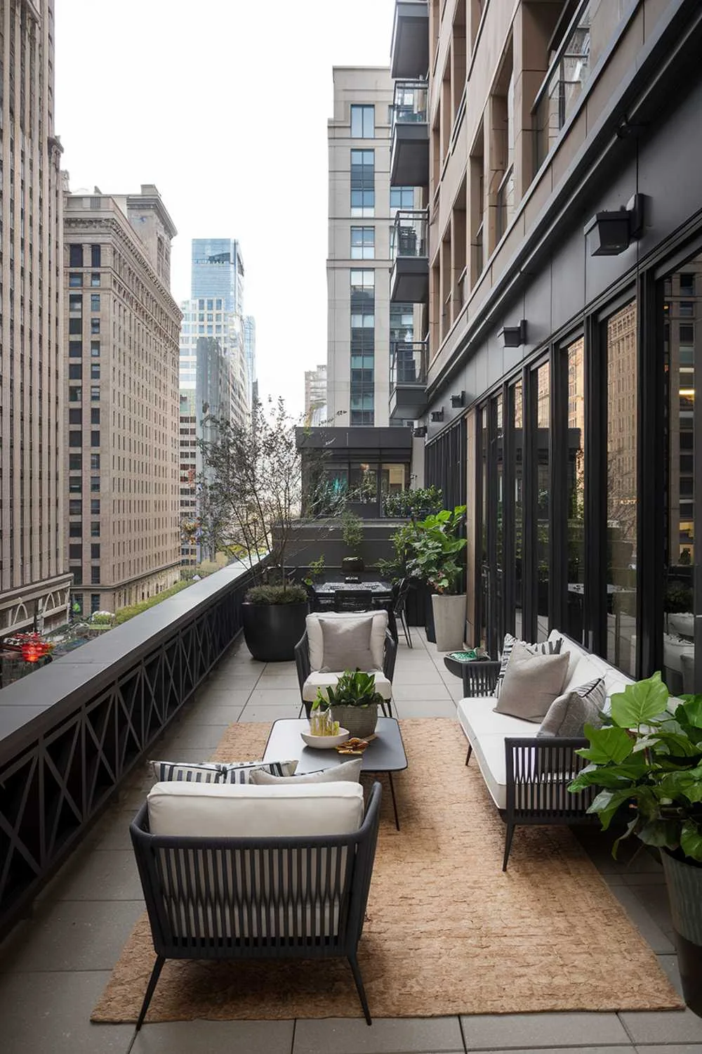 A large balcony with a stylish design and decor. The balcony has a dark wooden railing and is furnished with a white couch, a few potted plants, and a table. There is a beige rug on the floor. The background reveals a cityscape with tall buildings.