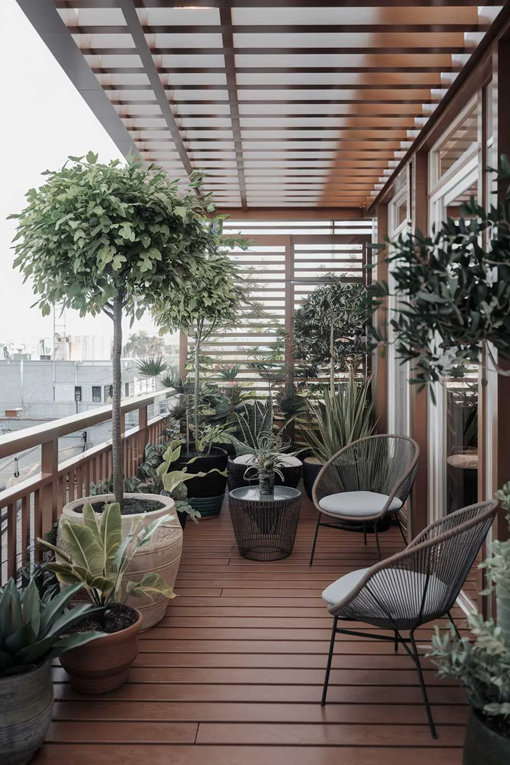 A stylish large balcony design and decor. There is a wooden deck with railing and a few chairs. There are potted plants of various sizes, including large trees. There is a small table between the chairs. The balcony is partially covered by a structure with a wooden slat design. The background reveals a cityscape with buildings.