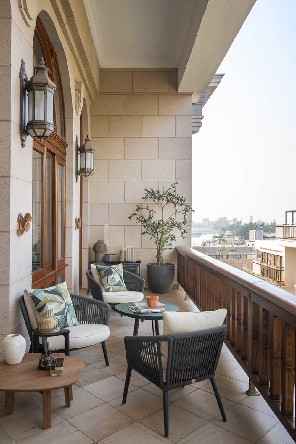 A large balcony with a stylish design and decor. The balcony has a wooden railing, a few chairs, and a small table. There's a potted plant in the corner. The balcony has a beige wall with a few decorative items like a lantern and a vase. The floor is made of tiles. The background reveals a city with buildings.