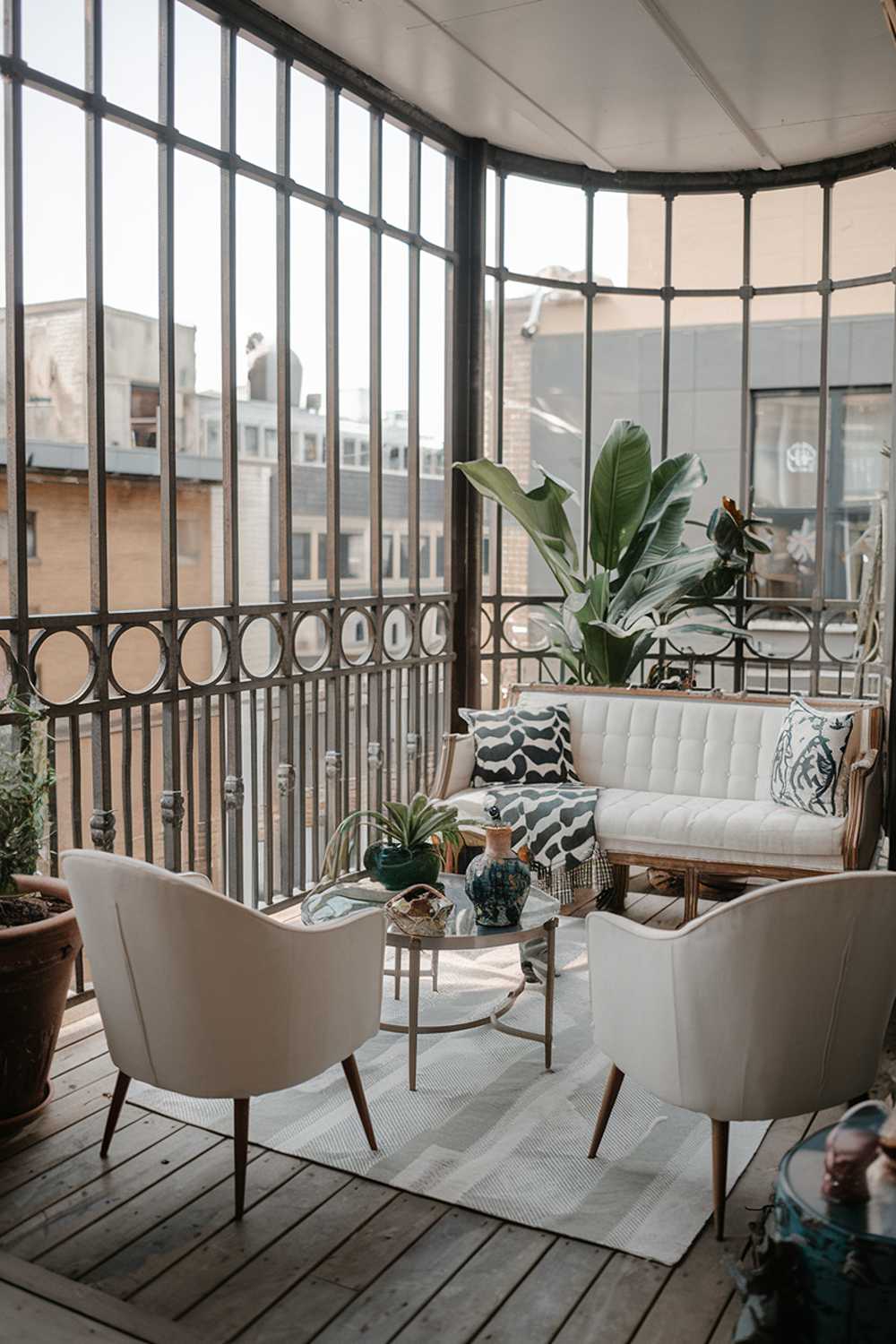 A balcony with a mix of modern and vintage decor. There's a white sofa, two white chairs, and a small table. On the table, there's a green plant and a few decorative items. The railing is made of metal with round bars. The floor is made of wood. There's a large potted plant near the sofa. The background reveals a cityscape with buildings.