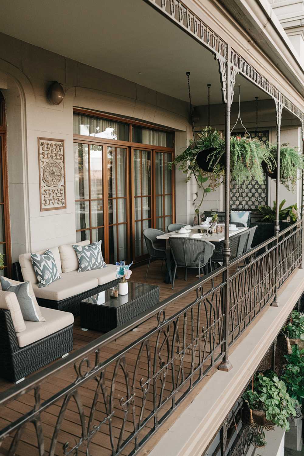 A large, stylish balcony with a comfortable seating area and a dining set. There's a coffee table between the seating and dining areas. The balcony railing is made of metal and has an intricate design. The floor is made of wooden planks. There are hanging plants near the railing. The wall has a decorative panel. The overall setup is perfect for relaxation and socializing.