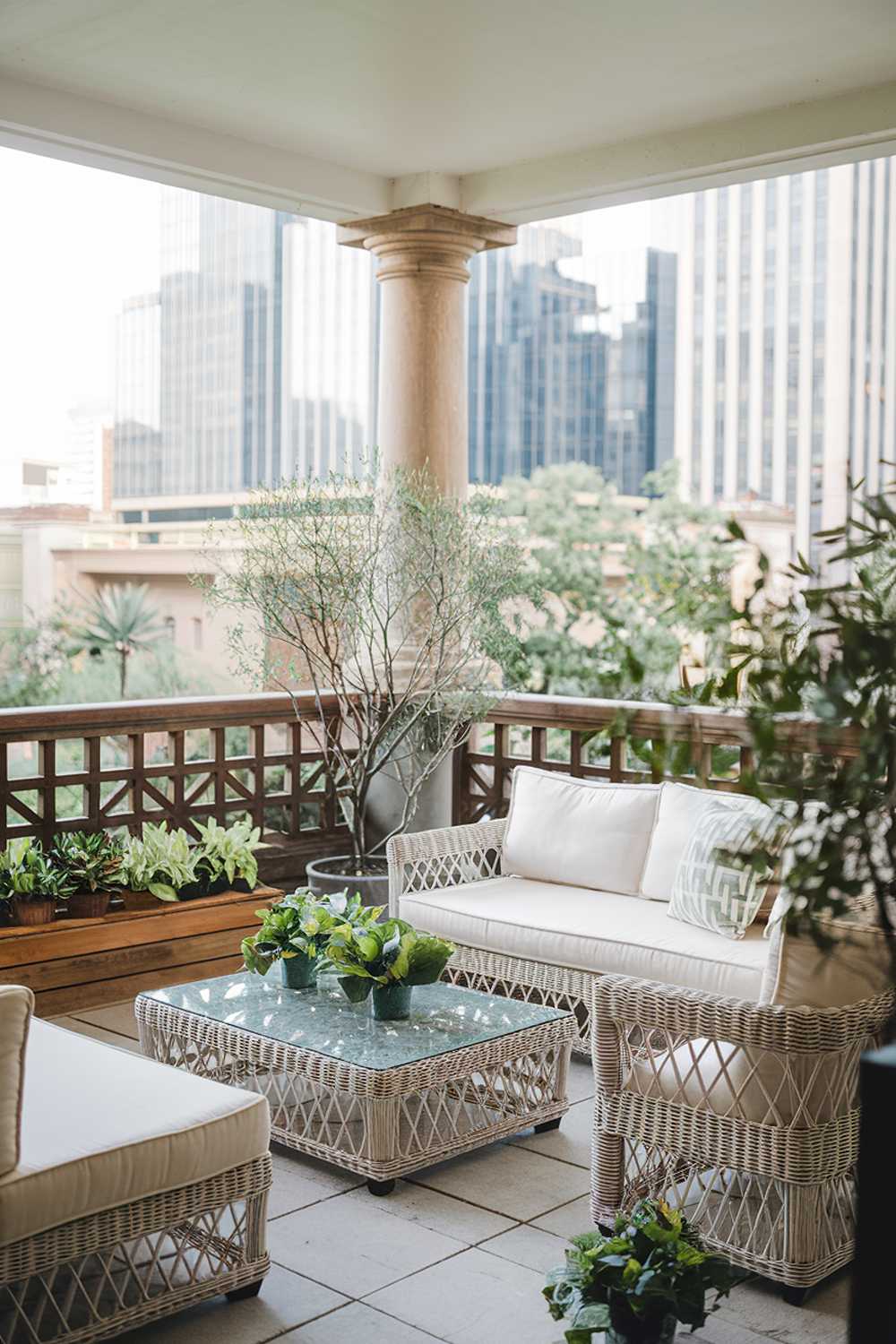 A large balcony with stylish decor. The balcony has a wooden railing and is furnished with a white wicker sofa, a coffee table made of the same material, and a few green potted plants. There's a wooden box beside the sofa. The floor is made of tiles. The background reveals a beautiful view of a city with tall buildings. The overall ambiance of the photo is serene and inviting.