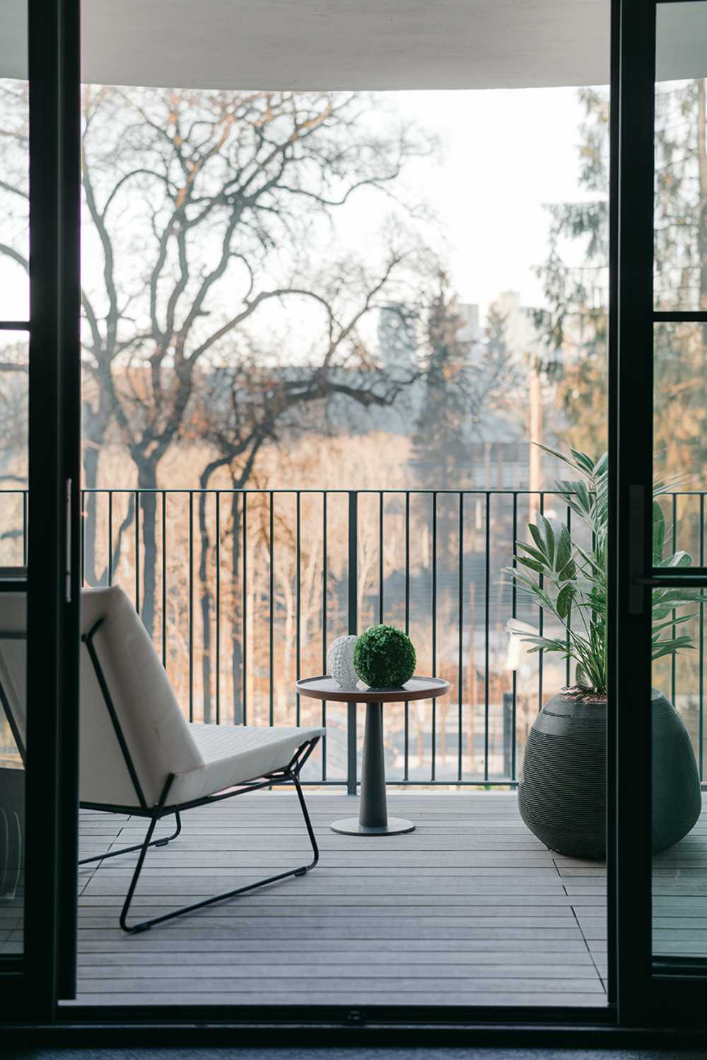 A high-end modern balcony with a stylish design and decor. There's a white lounge chair and a small round table. On the table, there's a green plant and a white decorative object. The balcony has a railing made of black metal. The floor is made of wooden planks. The background reveals a serene landscape with trees and buildings.