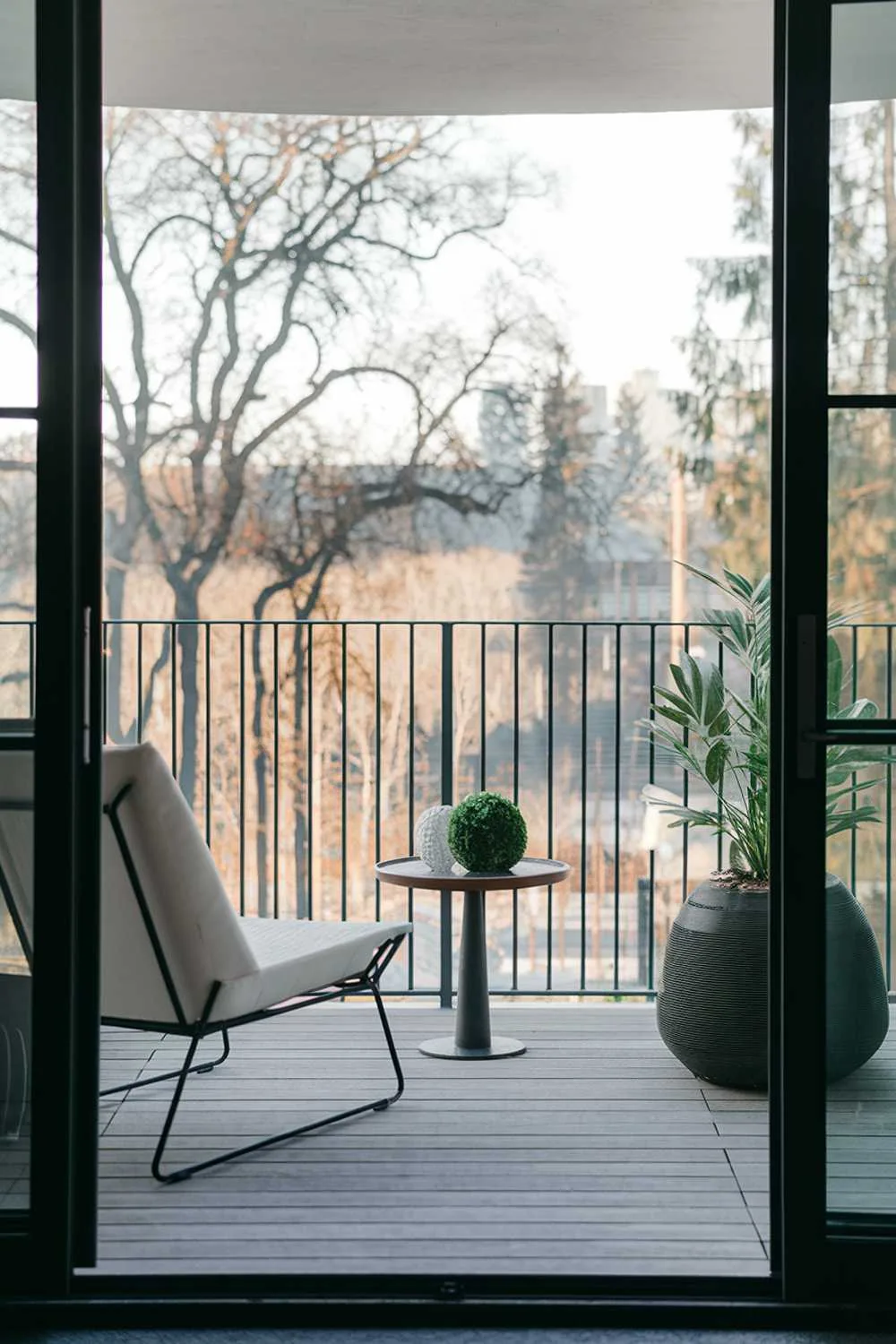 24 Must-See Modern Balcony Design Ideas for Stylish Living 3 A high-end modern balcony with a stylish design and decor. There's a white lounge chair and a small round table. On the table, there's a green plant and a white decorative object. The balcony has a railing made of black metal. The floor is made of wooden planks. The background reveals a serene landscape with trees and buildings.