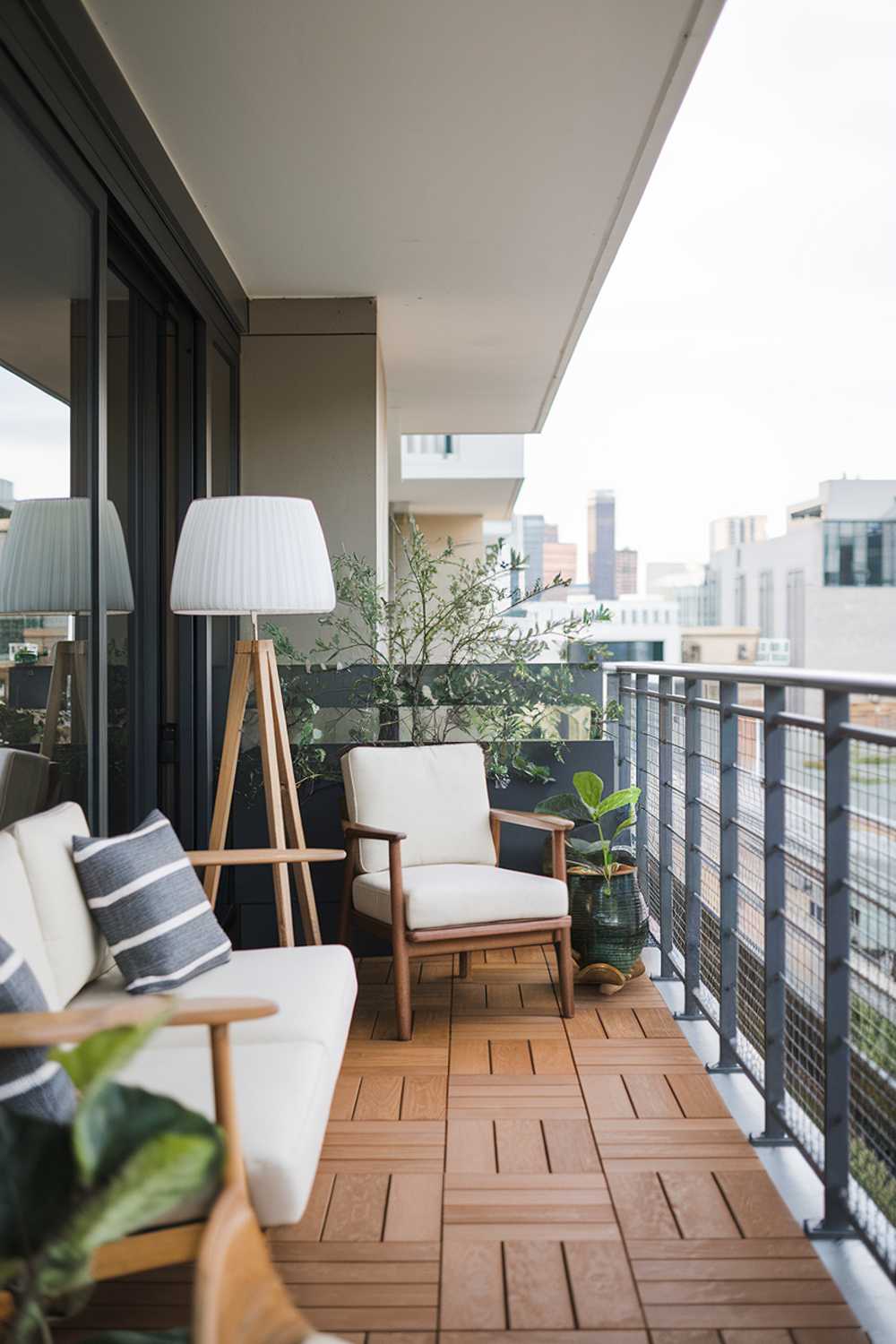 A modern balcony with stylish decor. The balcony has a wooden floor. There's a white couch, a wooden chair, a white lampshade, and a green plant. The railing is made of metal. The background reveals a cityscape with buildings.