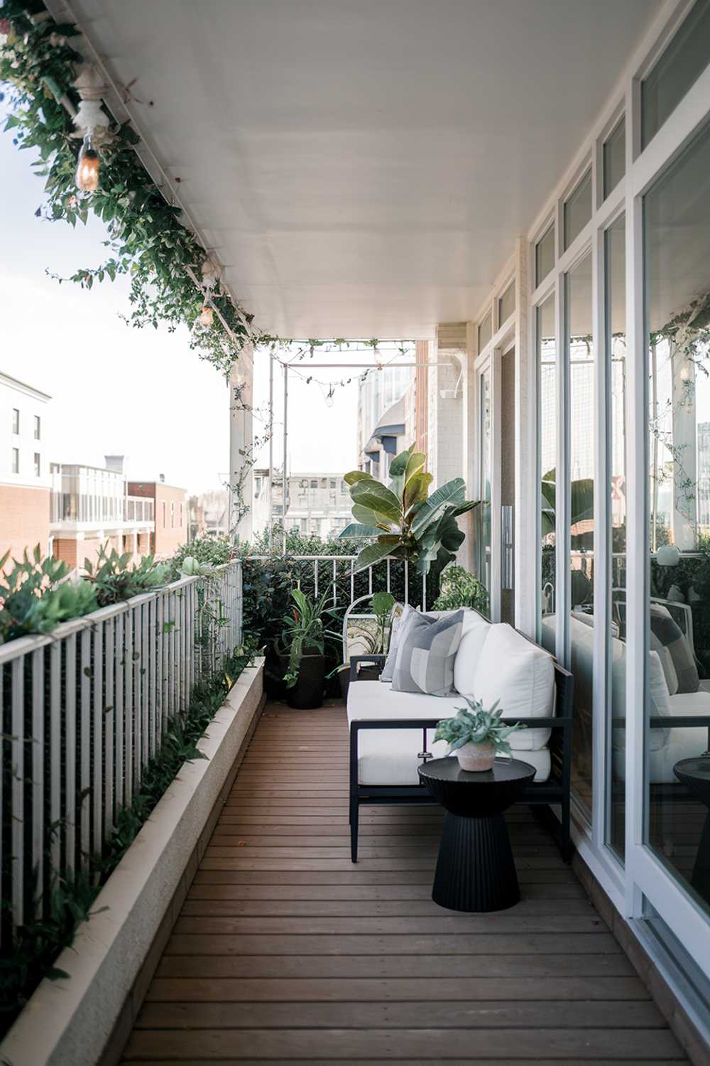 A modern balcony with a stylish design and decor. The balcony has a white railing and is surrounded by greenery. The floor is made of wooden planks. There is a white couch, a black side table, and a potted plant on the balcony. The background reveals a cityscape with buildings.