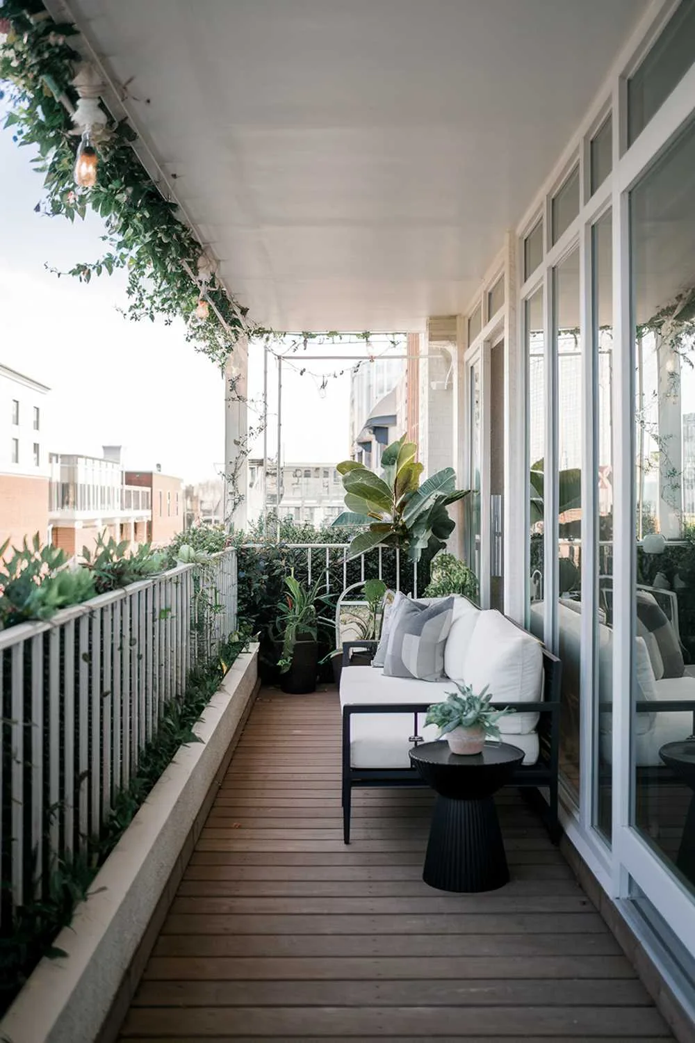 24 Must-See Modern Balcony Design Ideas for Stylish Living 5 A modern balcony with a stylish design and decor. The balcony has a white railing and is surrounded by greenery. The floor is made of wooden planks. There is a white couch, a black side table, and a potted plant on the balcony. The background reveals a cityscape with buildings.