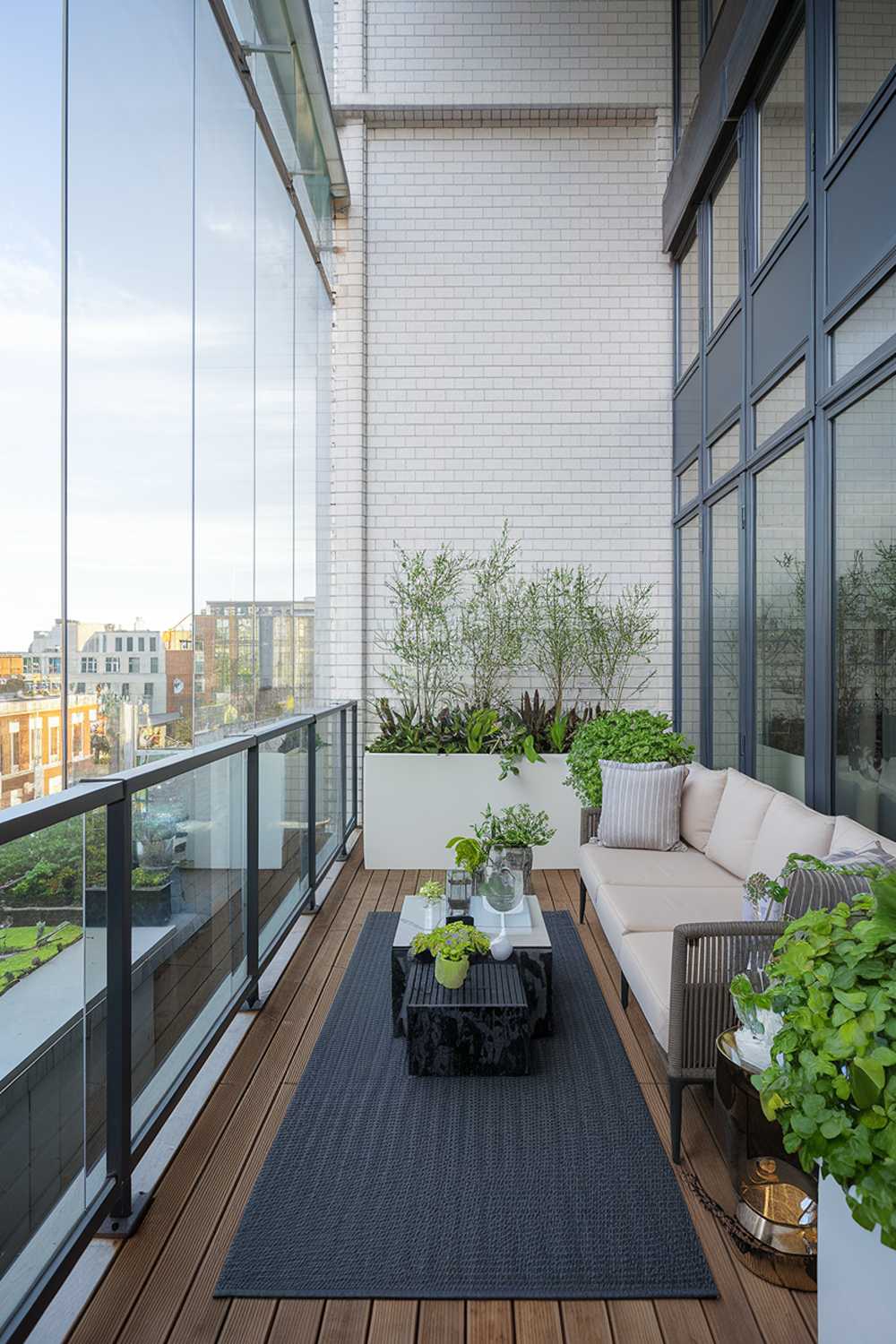 A modern balcony with a highly detailed design. The balcony has a glass railing and is made of wood. There's a beige sofa, a white planter, and a black outdoor rug on the balcony. The wall behind the balcony has a white brick pattern. There are green plants on the balcony. The view outside the balcony has a cityscape with buildings.