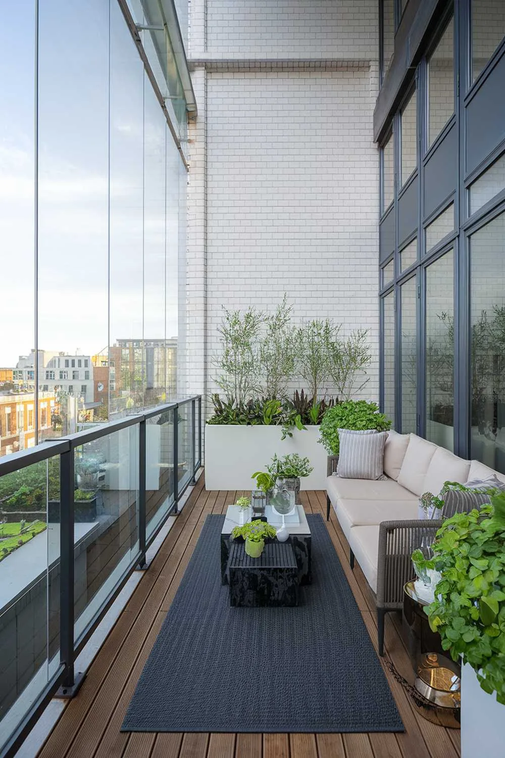 24 Must-See Modern Balcony Design Ideas for Stylish Living 12 A modern balcony with a highly detailed design. The balcony has a glass railing and is made of wood. There's a beige sofa, a white planter, and a black outdoor rug on the balcony. The wall behind the balcony has a white brick pattern. There are green plants on the balcony. The view outside the balcony has a cityscape with buildings.