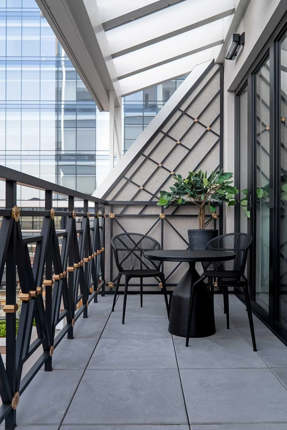 24 Must-See Modern Balcony Design Ideas for Stylish Living 14 A modern balcony design and decor with a highly detailed railing. The railing has a geometric pattern with black metal bars and gold metal connectors. The floor is made of large grey tiles. There's a small round black table and two black chairs on the balcony. A potted plant with green leaves is placed on the table. The wall behind the railing has a textured surface. The balcony is partially covered, with a white structure above it. The background reveals a modern building with a glass facade.