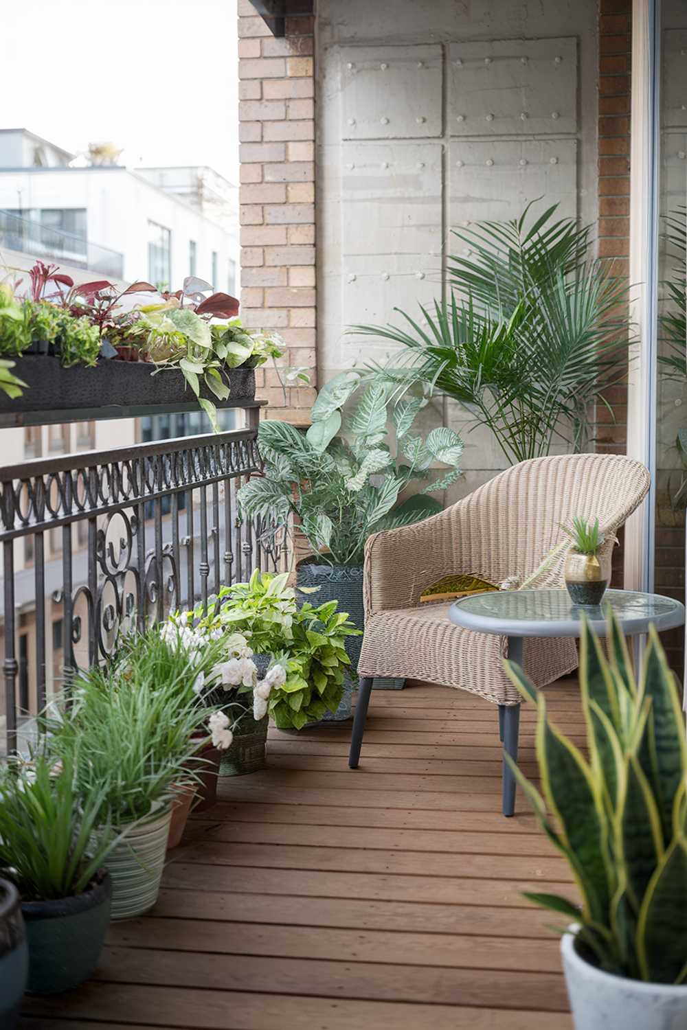 A modern balcony design and decor. The balcony has a wooden floor and is adorned with a variety of plants in pots. There's a wicker chair and a small table. A potted plant is placed beside the chair. The railing is made of metal and has a decorative pattern. The background reveals a cityscape with buildings.