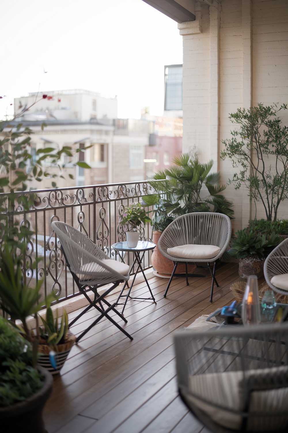 A modern balcony with a detailed design and decor. The balcony features a wooden floor, a few chairs and a table, and a few potted plants. The railing is made of iron and has a decorative design. The background reveals a cityscape with buildings. The overall ambiance is serene and inviting.