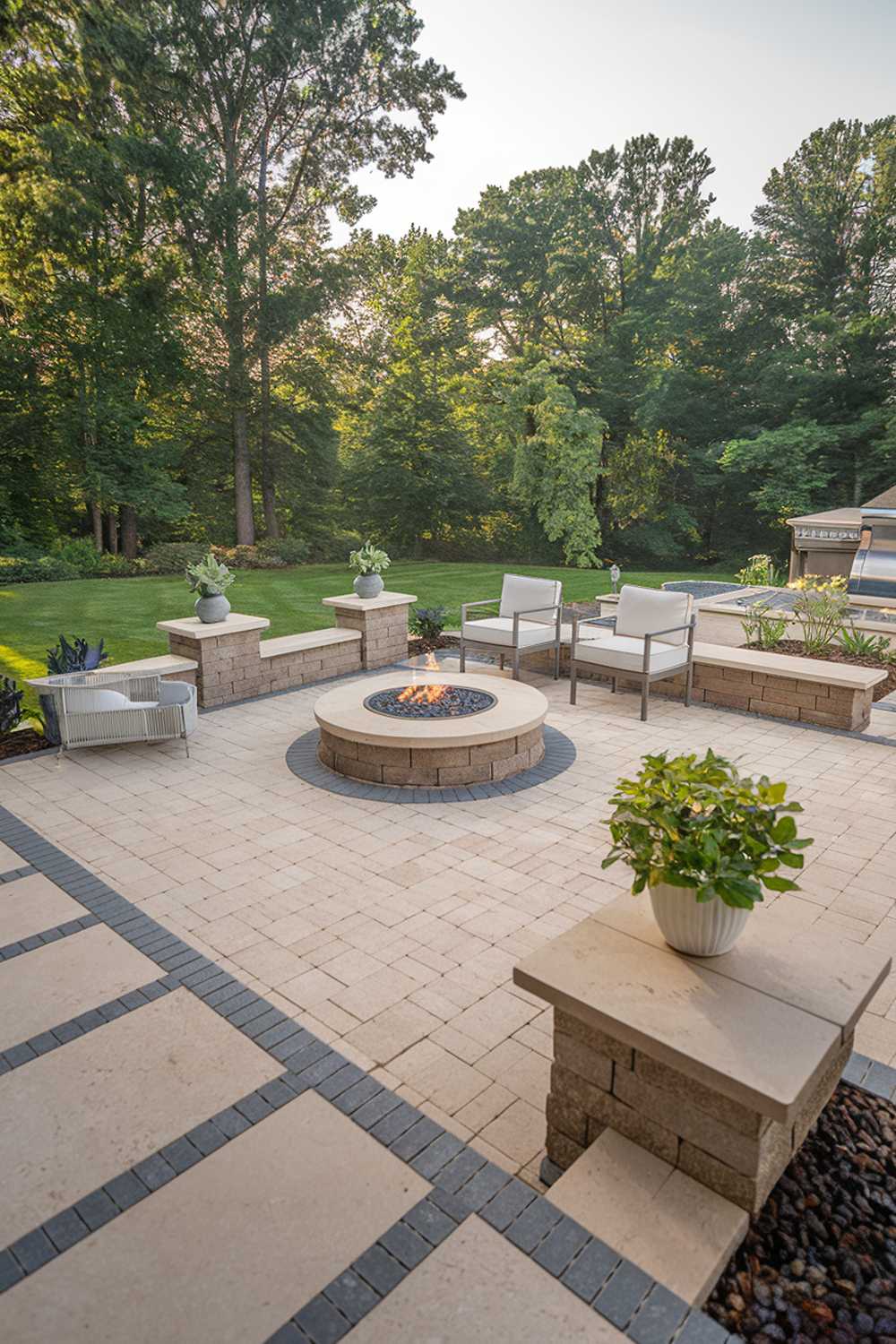 A paver patio design and decor. The patio has a rectangular shape and is made of beige and grey pavers. There is a round fire pit in the middle of the patio, surrounded by beige pavers. There are two white chairs and a white table near the fire pit. There is a green plant in a white pot near the fire pit. The background is a lush green garden with tall trees.