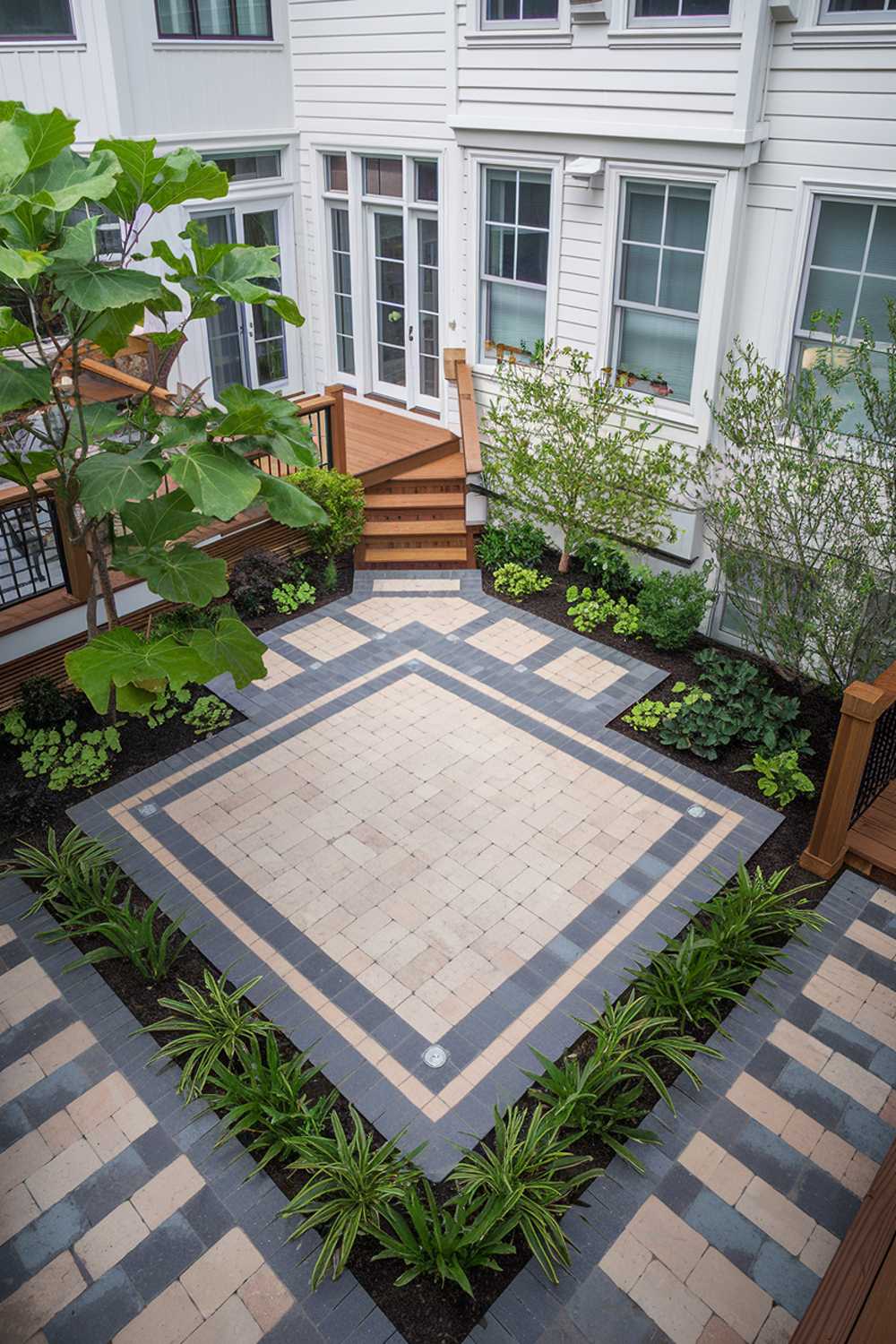 A paver patio design with a mix of grey and beige pavers. The patio has a square shape and is surrounded by lush green plants. There is a wooden deck connected to the patio, leading to a set of stairs. The background reveals a white house with multiple windows.