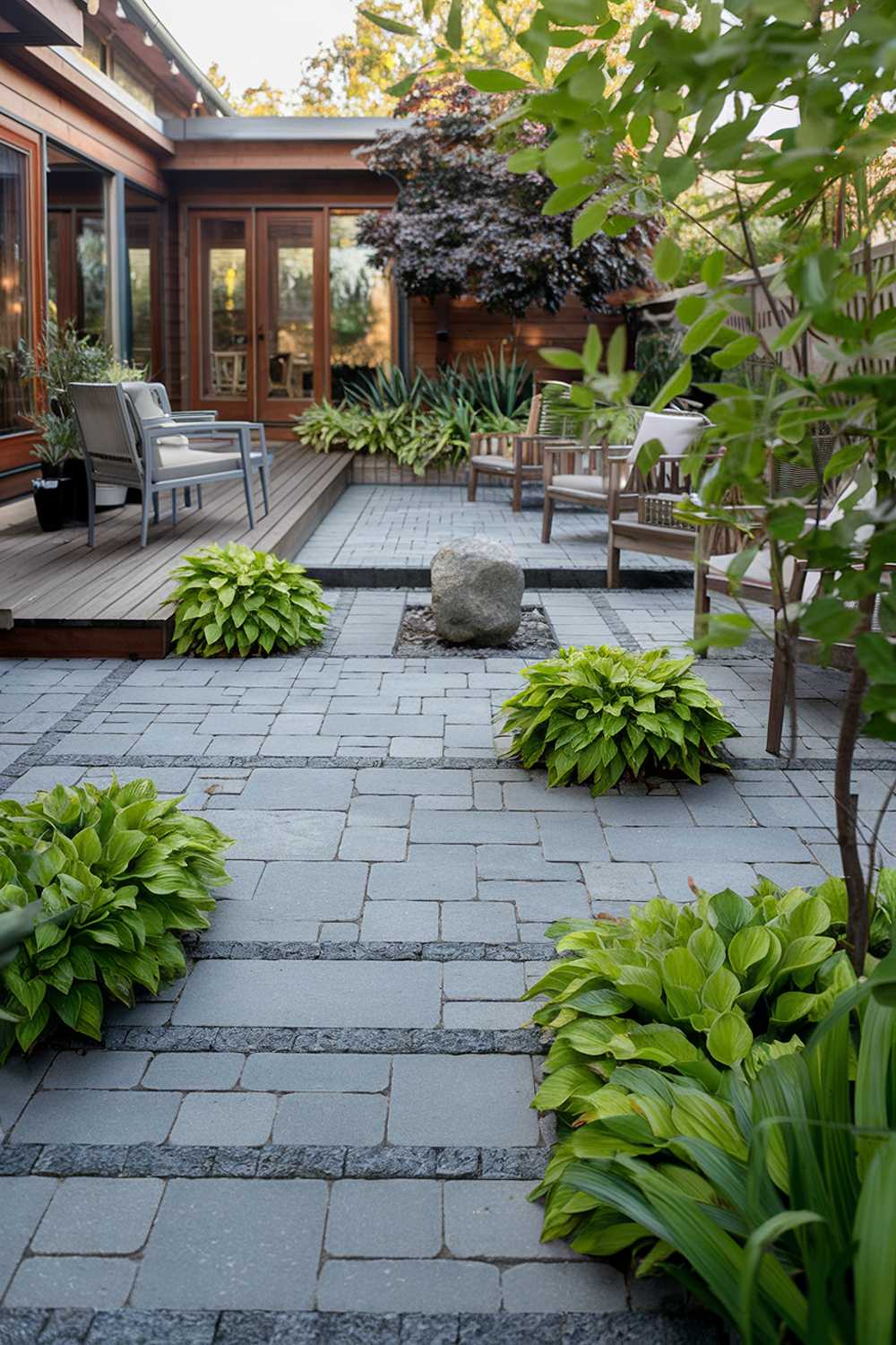 A paver patio design with a highly detailed stylish layout. The patio is made of grey pavers and is surrounded by lush green plants. There's a wooden deck with a few chairs and a table. A stone feature is placed in the middle of the patio. The background reveals a house with a wooden facade. The overall ambiance is serene and inviting.