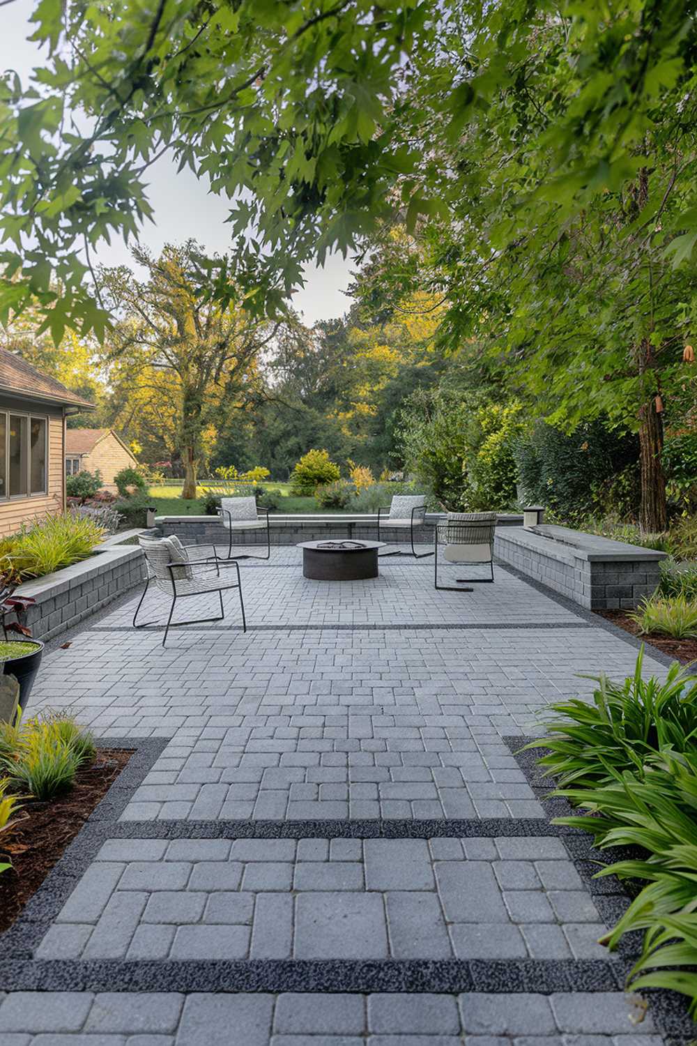 A backyard with a modern paver patio design. The patio is made of grey pavers and has a few chairs. There is a fire pit at one end of the patio. The patio is surrounded by lush greenery. The background contains a house and trees.