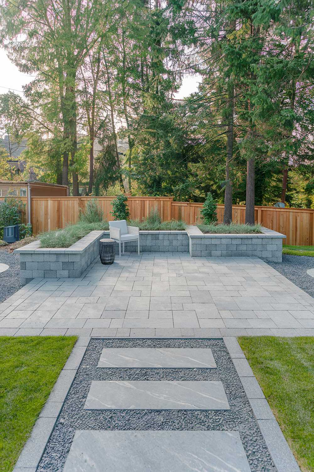 A modern paver patio design in a backyard. The patio is made of large, rectangular pavers in a medium grey hue. There are two paver paths leading to the patio from the grassy areas. The patio has a built-in planter in one corner, filled with greenery. There is a white outdoor chair and a small table on the patio. The background reveals a wooden fence and tall trees. The ground around the patio is covered with gravel.