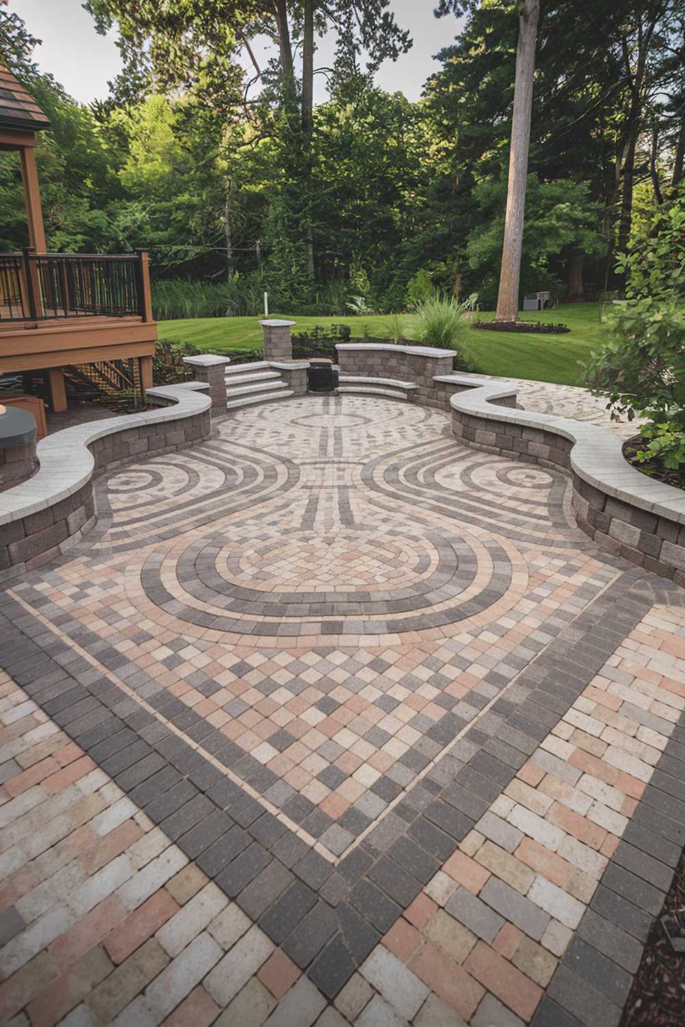 A backyard with a paver patio design. The patio has a square layout with curved edges and is made of interlocking pavers in a mix of grey, beige, and brown tones. The pavers form a pattern with circles and waves. The background contains a wooden deck, lush greenery, and tall trees.