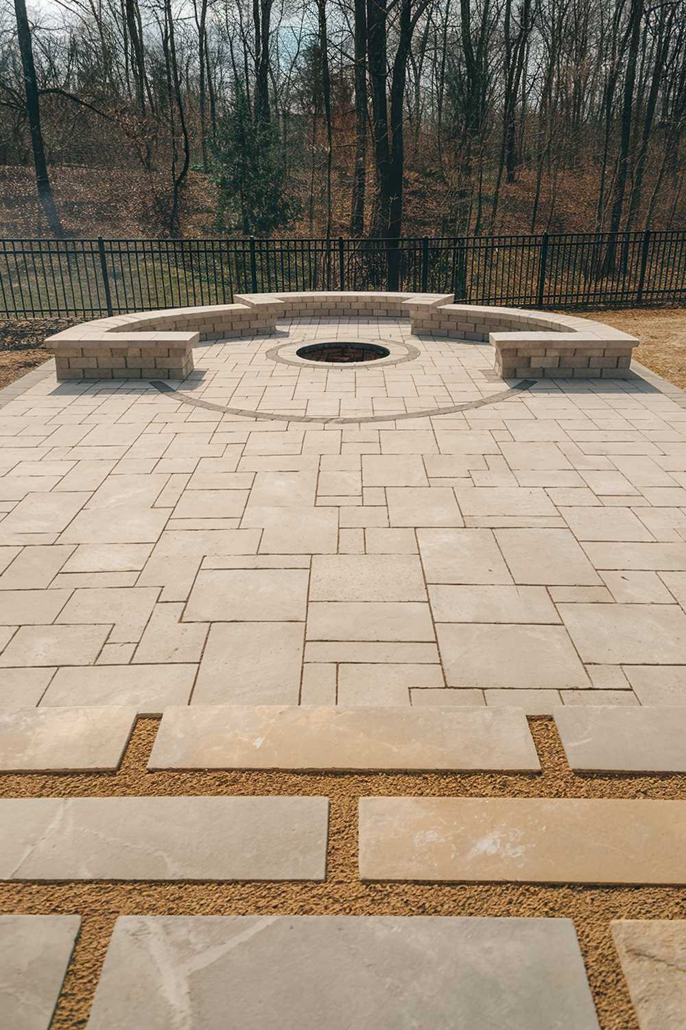 A backyard with a modern paver patio design. The patio is made of large, rectangular pavers in a soft beige hue. A few pavers are placed in a circular pattern, creating a space for a table or chairs. The background contains a fence and trees. The ground is covered with a layer of light brown gravel.