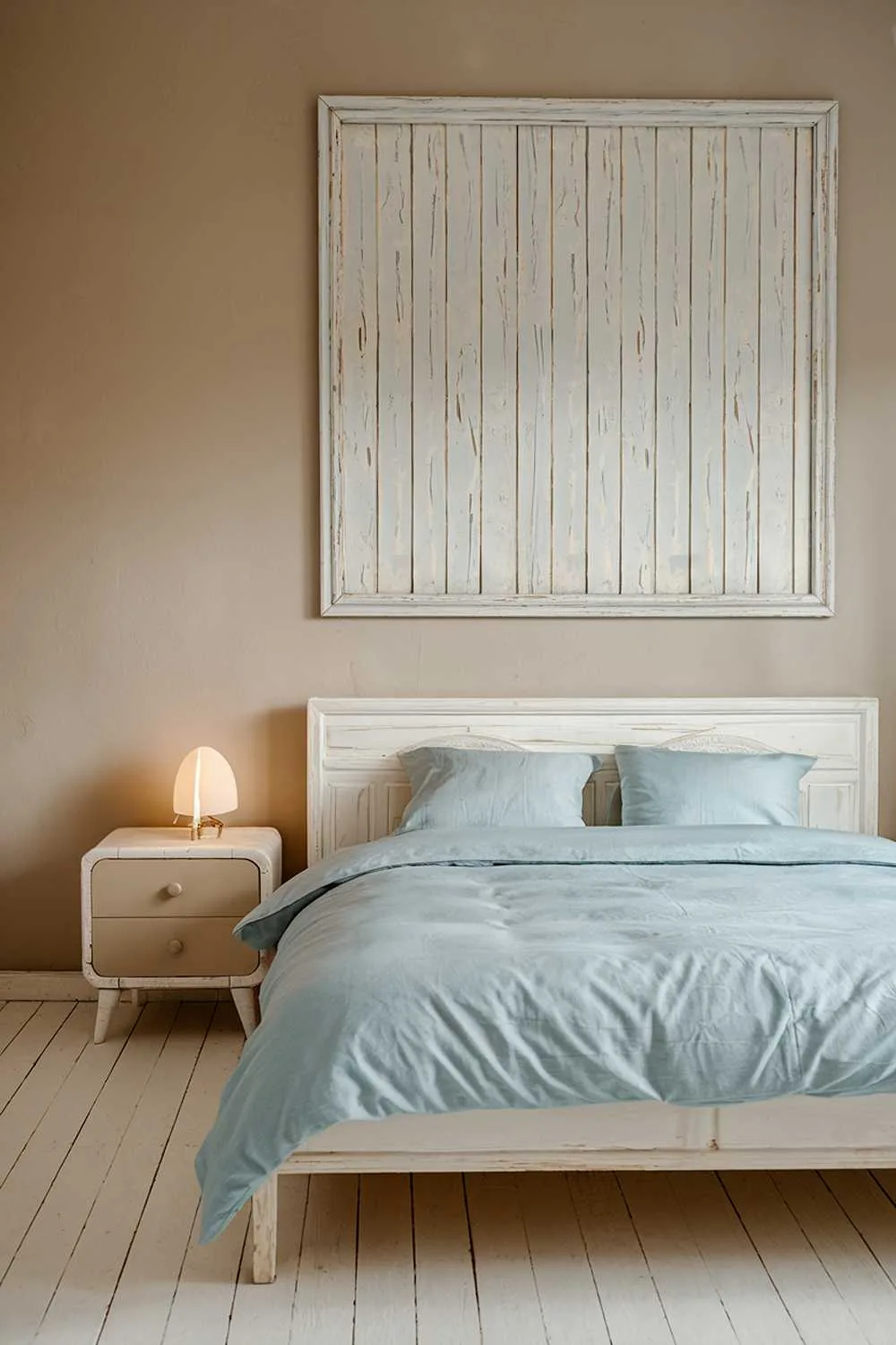 21 Scandi Bedroom Ideas for a Cozy, Minimalist Retreat 1 A Scandi bedroom decor. The room has a white wooden floor and beige walls. There is a white wooden bed with a light blue duvet and two white pillows. Next to the bed, there is a white wooden nightstand with a beige accent and a small white lamp. On the wall above the bed, there is a large white wooden frame. The room has a soft, warm lighting.