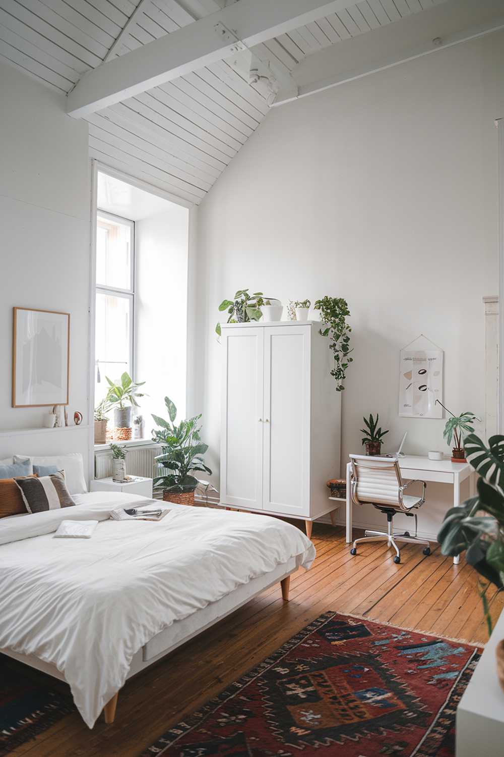 A Scandi bedroom with a high ceiling. The room has white walls, wooden floors, and a few large, white furniture pieces. There's a double bed with a white duvet and a few pillows. Next to the bed is a white nightstand. On the other side of the room, there's a white wardrobe and a white desk with a chair. The room has a few potted plants and a rug. The lighting is soft.