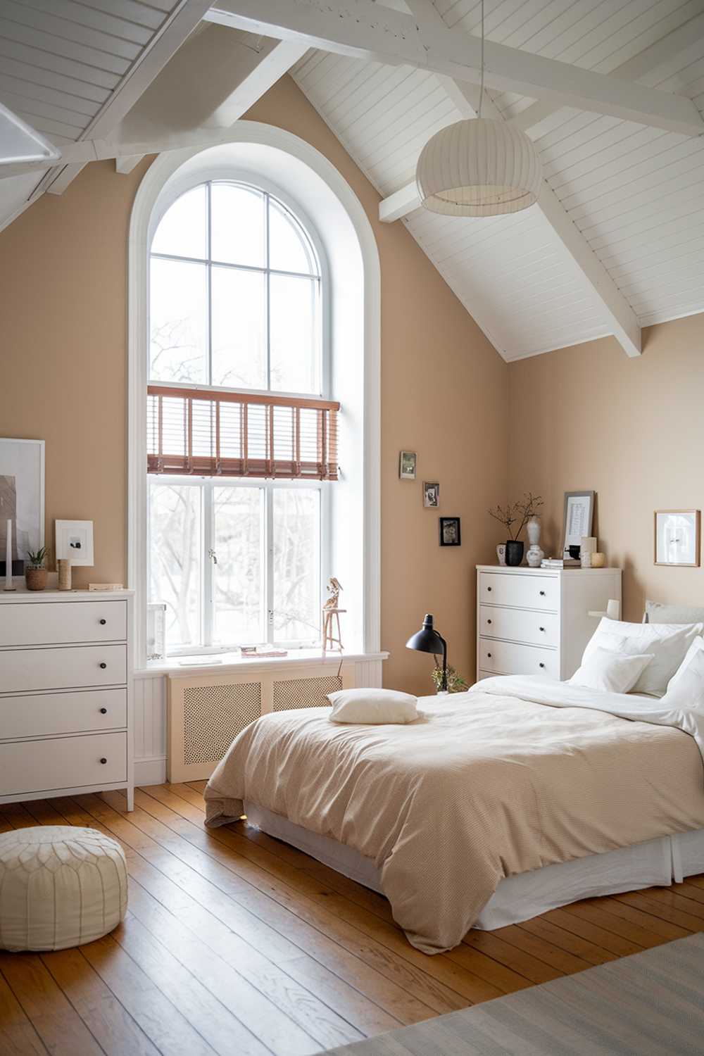 A Scandinavian bedroom with a high ceiling. The room has a large window with wooden blinds that lets in natural light. There's a queen-sized bed with a beige duvet and white pillows. The walls are painted in a soft beige hue. The floor is made of oak wood. There's a white IKEA Malm dresser with 5 drawers next to the bed. The room has a white pouf near the bed. The room also has a white lampshade with a wooden base.