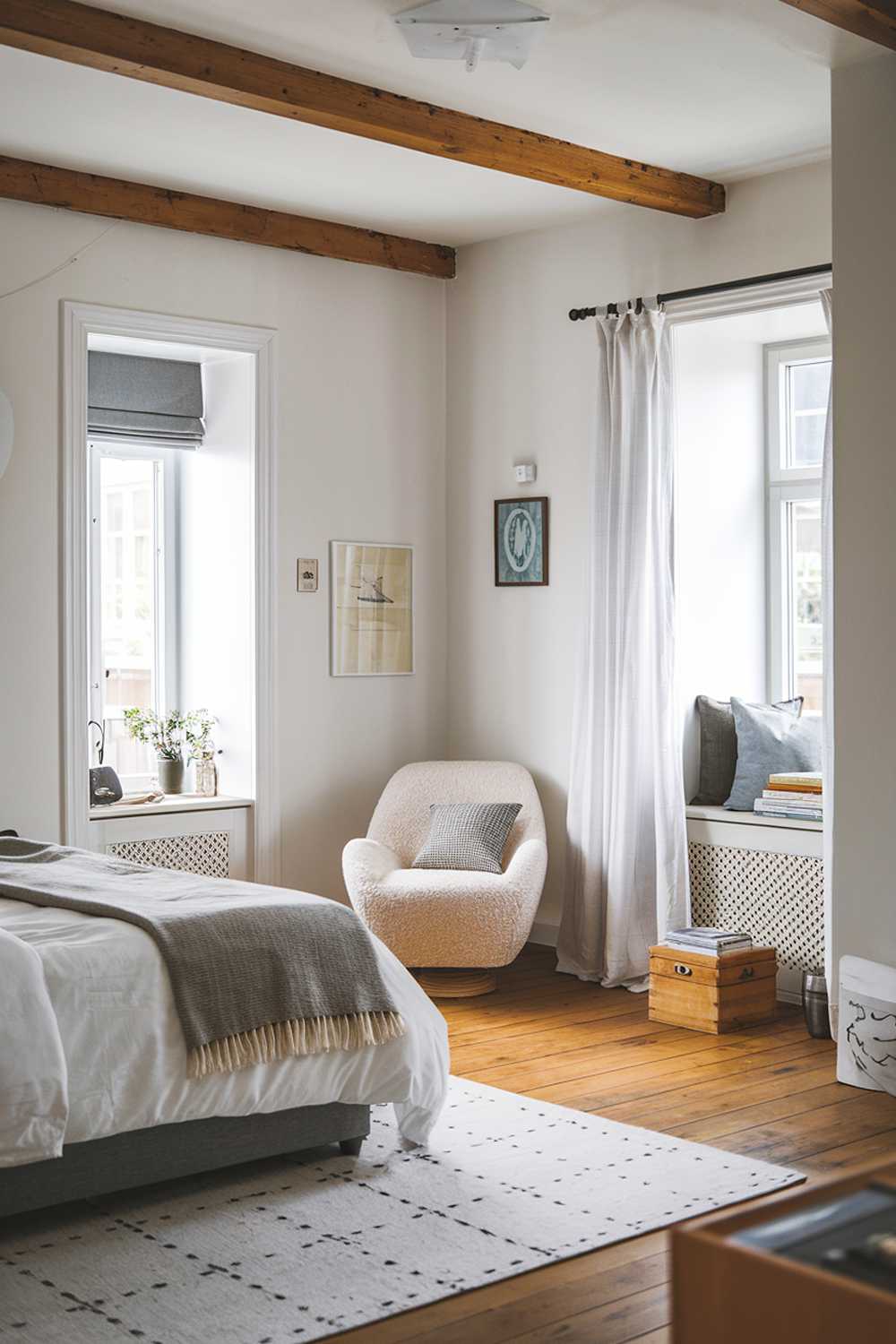 A Scandinavian bedroom with stylish decor. The room has a white walls, a wooden floor, and a wooden beam ceiling. There's a cozy chair in the corner near the window. The bed is made with white sheets and a gray blanket. There's a gray chair at the foot of the bed. The room has a few decorative items, including a wooden box and a few books. The window has a white curtain.