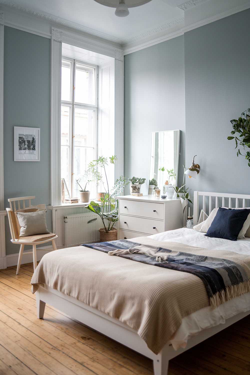 A Scandinavian bedroom decor. The room has a white wooden bed frame with a beige blanket and a dark blue throw pillow. There's a white chair with a beige cushion next to the bed. A white dresser with a mirror is placed near the bed. The walls are painted in light blue. There's a small plant near the dresser. The room has a wooden floor.