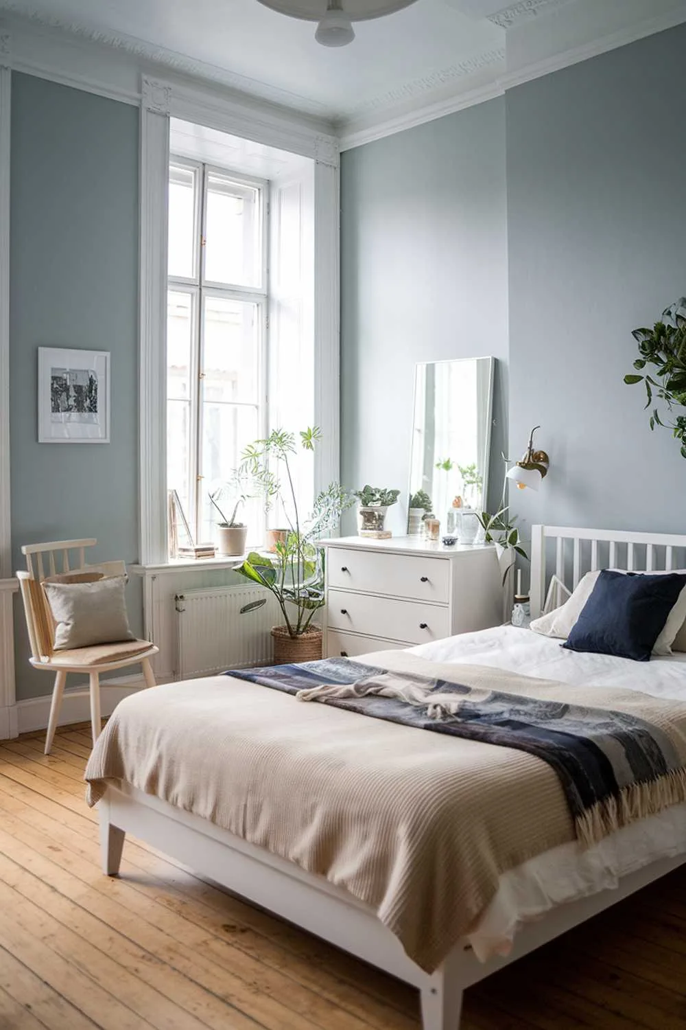 21 Scandi Bedroom Ideas for a Cozy, Minimalist Retreat 13 A Scandinavian bedroom decor. The room has a white wooden bed frame with a beige blanket and a dark blue throw pillow. There's a white chair with a beige cushion next to the bed. A white dresser with a mirror is placed near the bed. The walls are painted in light blue. There's a small plant near the dresser. The room has a wooden floor.
