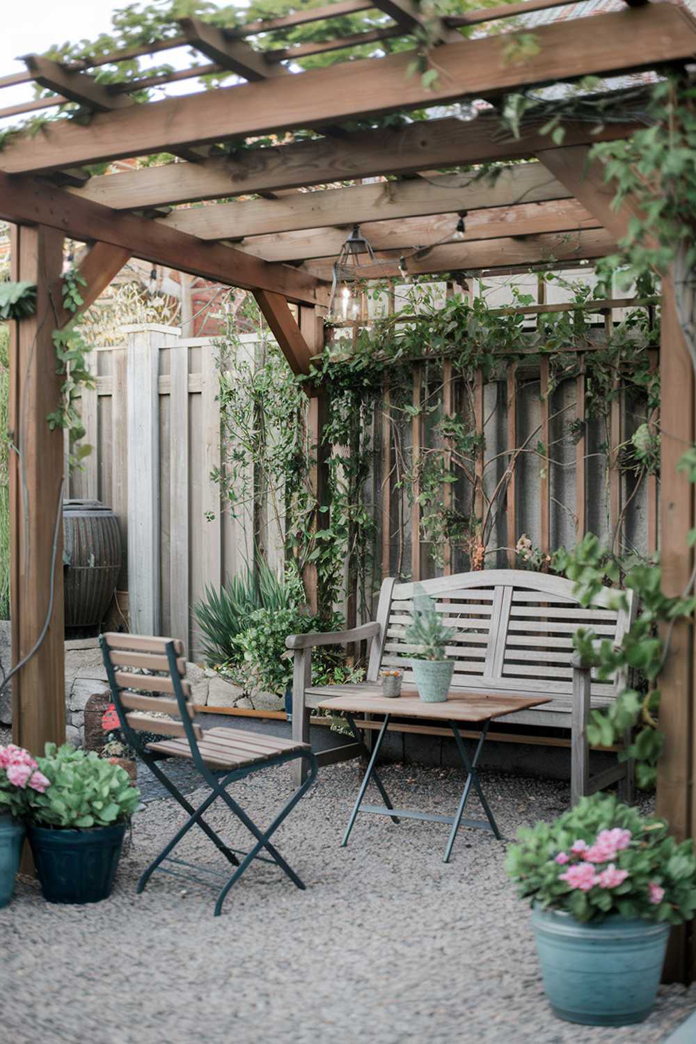 A small patio design and decor. There is a wooden pergola with climbing plants. A wooden bench and a few chairs are placed around a small table. The ground is covered with gravel. There are potted plants and a lantern hanging from the pergola. The background reveals a fence and a wall. The lighting is soft.