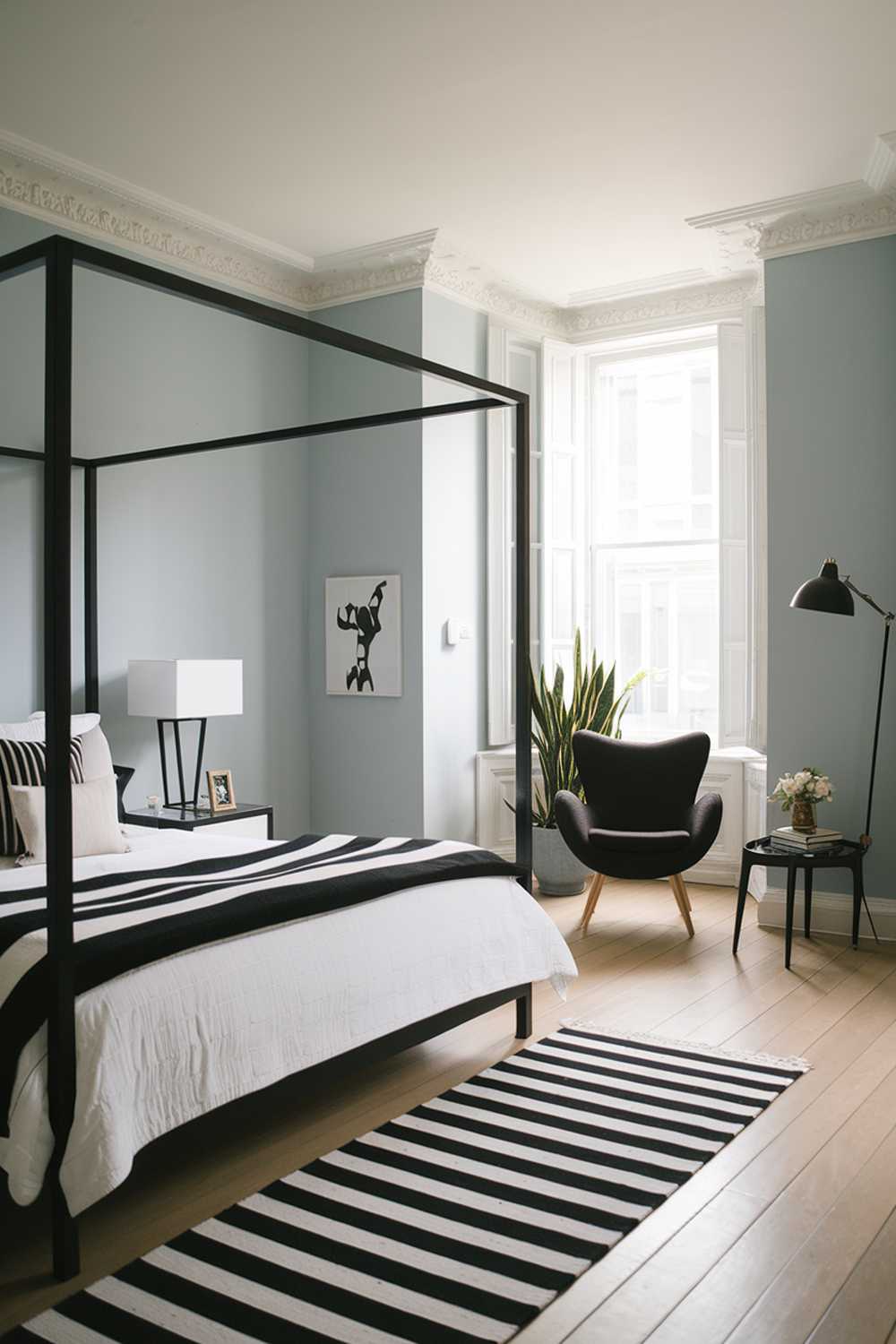 A stylish bedroom with a modern design and decor. The room has a white bed with a black frame and a black and white striped blanket. There is a black chair in the corner near the window. The walls are painted a light blue color. The flooring is a light wood. A white lamp is near the bed and a black lamp is near the chair. The room has a plant near the window.