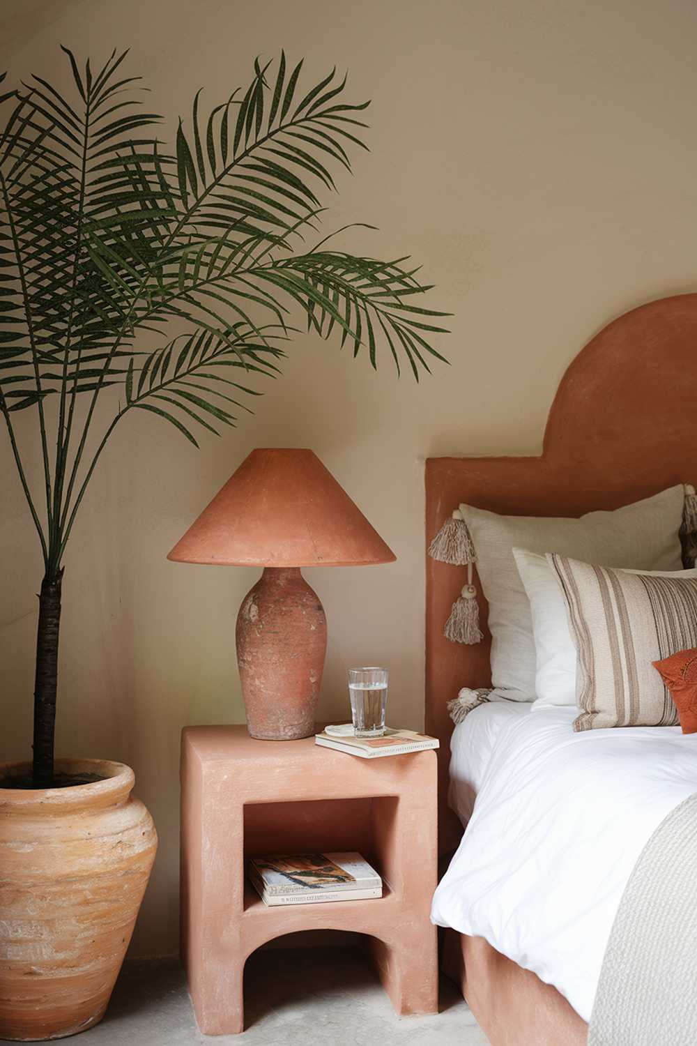 A terracotta bedroom design. The room features a comfortable bed with a white duvet and a variety of pillows. A terracotta lampshade is placed on the nightstand, which also holds a glass of water and a book. A terracotta pot with a fake palm tree stands near the bed. The walls are painted in a soft beige color.