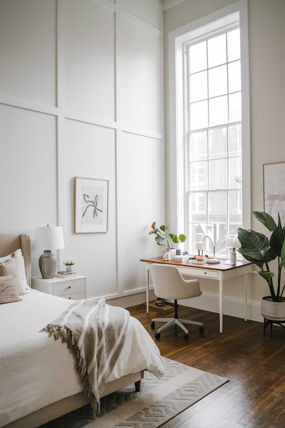 A white bedroom design and decor. The room has a high ceiling and is lit by a large window. The room has a bed, a nightstand, a desk, a chair, and a plant. The walls have simple white frames. The floor is wooden.