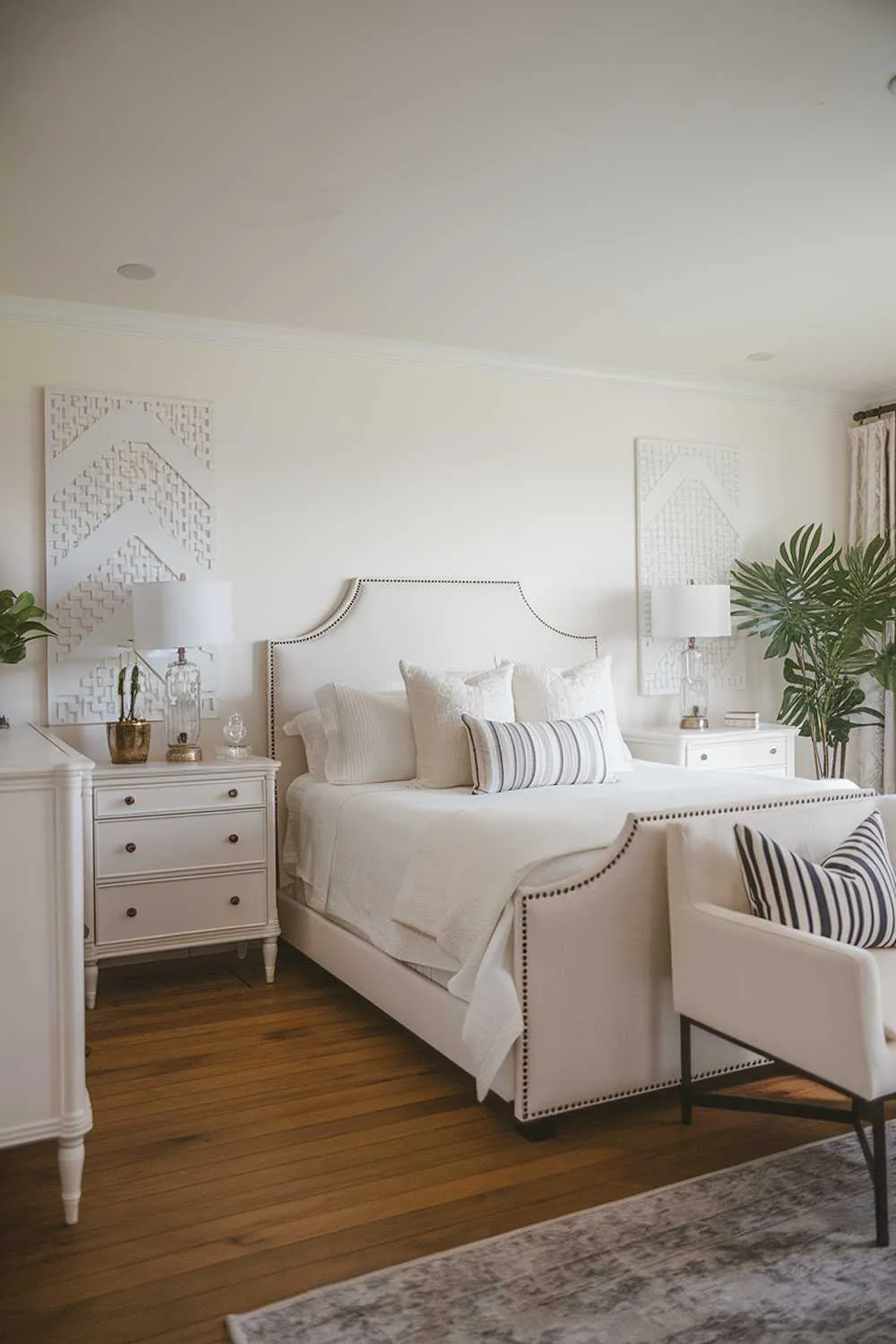 A cozy all white bedroom design and decor. The room features a queen-size bed with a white headboard. There's a white dresser, a nightstand, and a chair. The bed is placed in the middle of the room. The floor is made of hardwood. The lighting is soft. There's a potted plant near the nightstand. The walls are decorated with a few pieces of white artwork.